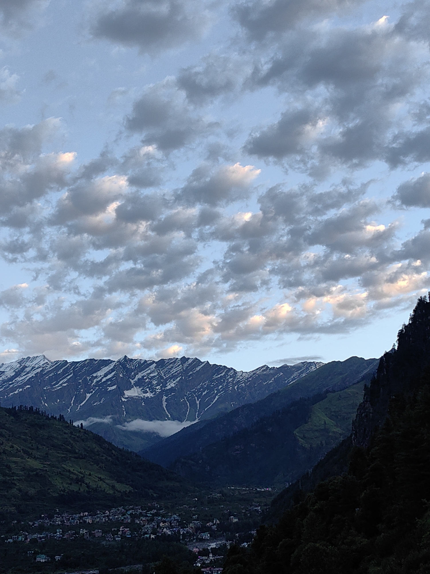 The image depicts a panoramic view of the mountainous region surrounding Vashisht, India. In the foreground, a dense, dark forest occupies the right side, contrasting with the lush green slopes of mountains that stretch across the middle ground. A small town, characterized by numerous buildings with colorful roofs, is nestled within the valley at the base of these slopes. Beyond the town, the majestic, snow-capped peaks of the Himalayas dominate the horizon, their rugged ridges sharply defined against the sky. The sky itself is a canvas of pale blue, adorned with a scattered array of white and grey clouds, some catching the soft light of either sunrise or sunset, casting a gentle glow. The scene is serene and natural, showcasing the dramatic landscape of Vashisht.