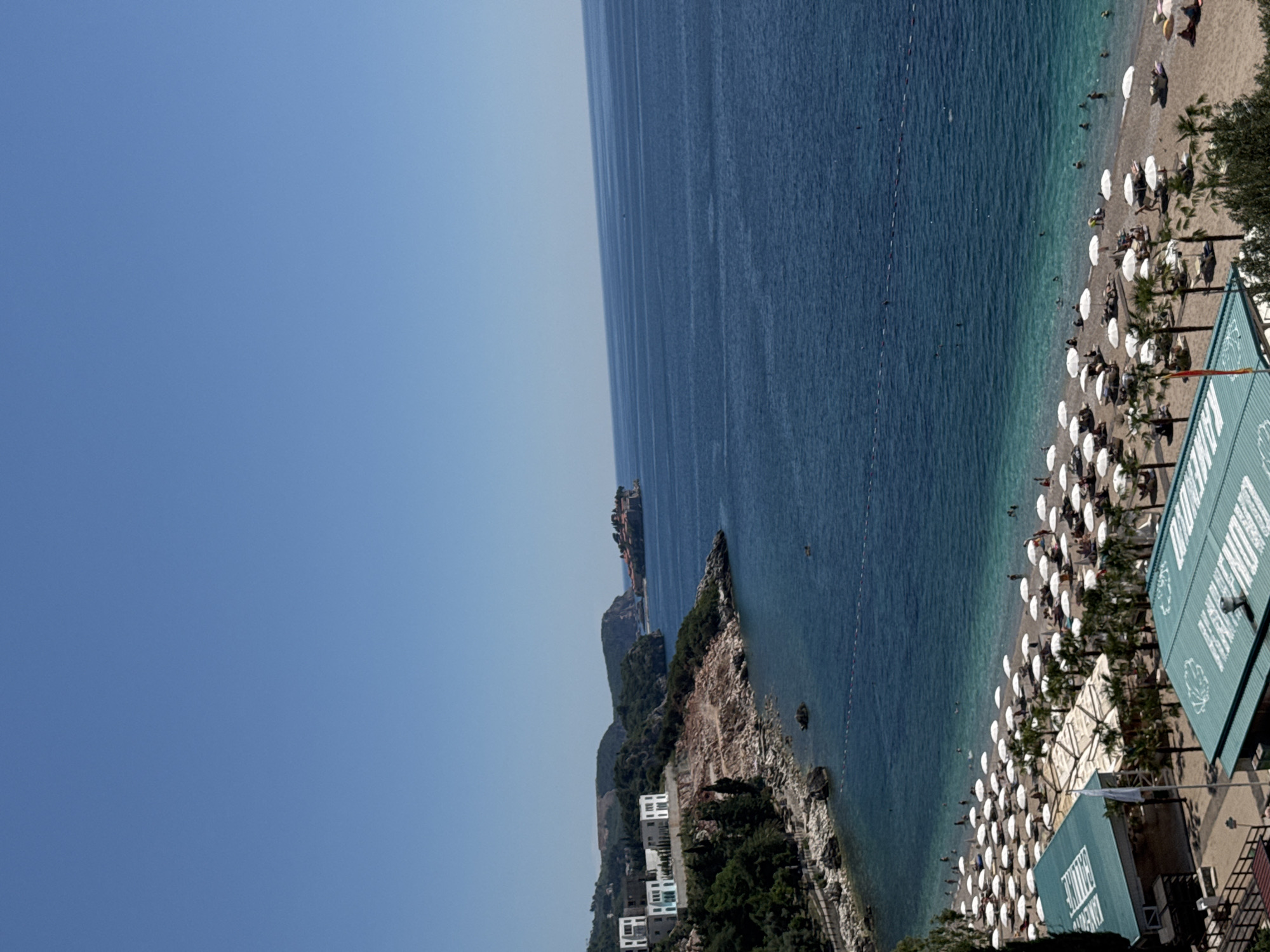 The scene is an aerial view of Kamenovo beach in Montenegro. The image captures a coastline with a mix of rocky and sandy beaches. The water is clear, varying from turquoise near the shore to deep blue further out. A dense line of white umbrellas dots the sandy beach, indicating a popular tourist destination. People are visible in the water and on the beach. Some small structures, possibly beach bars or restaurants, are also discernible. The sky is clear and blue, suggesting sunny weather. The view includes a stretch of the sea with an island visible in the distance. Further inland, there is a cliffside with sparse vegetation and some white buildings.
