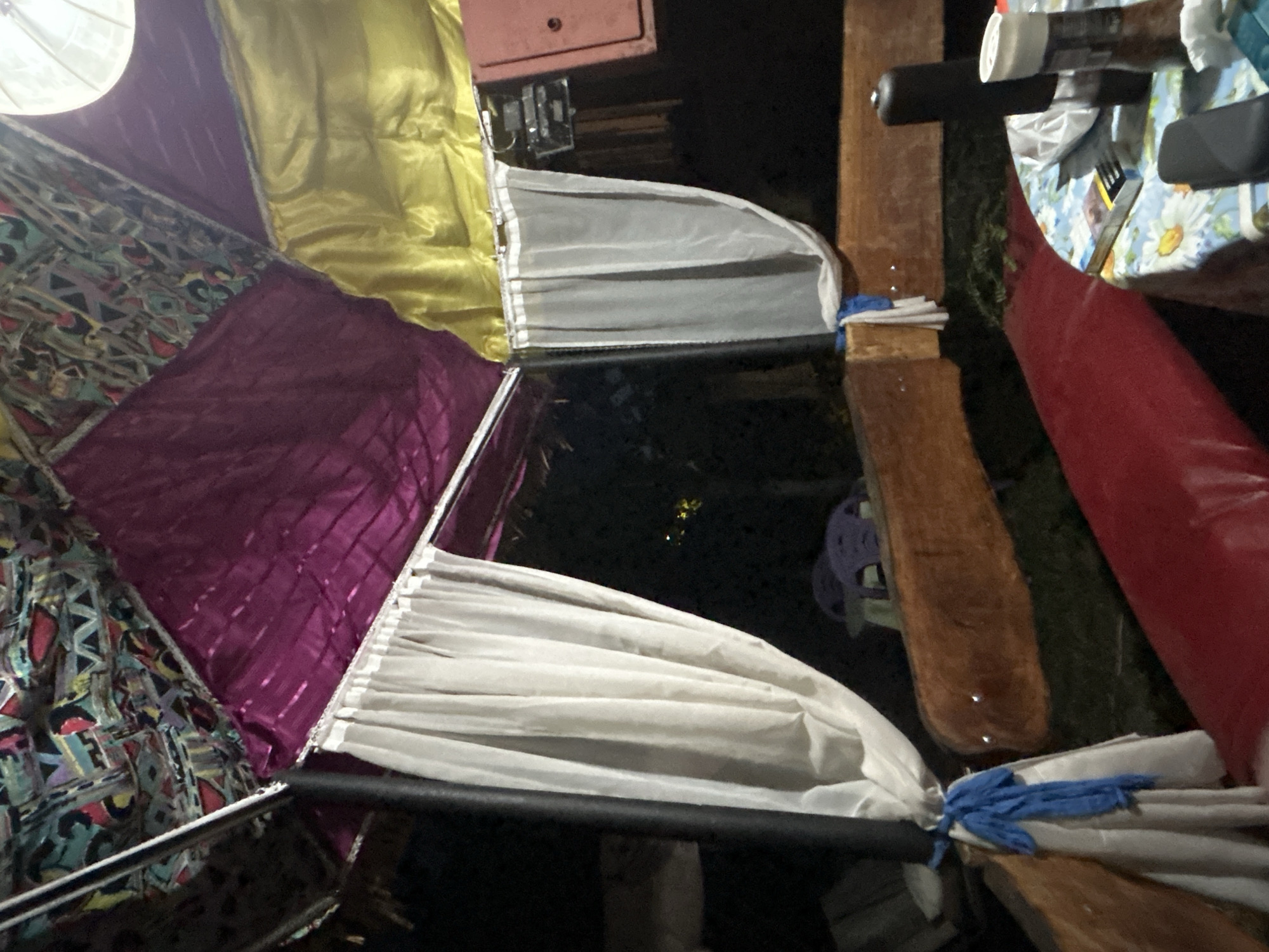 The image depicts the interior of a dimly lit, open-air structure, possibly a gazebo or a covered seating area, in Suhozem, Bulgaria. The ceiling is adorned with a patterned fabric and draped with vibrant purple and yellow materials. White curtains hang from the sides, partially obscuring the dark, outdoor environment beyond.

In the foreground, a round table covered with a floral tablecloth is visible. On the table are various items, including a pack of cigarettes, what appears to be a pepper grinder, and other unidentifiable objects. Two dark-colored seats or cushions are also present. A long, red cushioned bench runs along the lower part of the frame.

A wooden bench or railing with a curved edge is situated behind the red bench. A black pole, possibly a structural support, is visible on the left, with a white curtain tied to it with a blue rope. In the background, through the curtains, faint lights suggest the presence of other structures or lights in the distance. A portion of a purple chair is also visible in the middle ground. The overall impression is of a casual, outdoor relaxation or gathering space.
