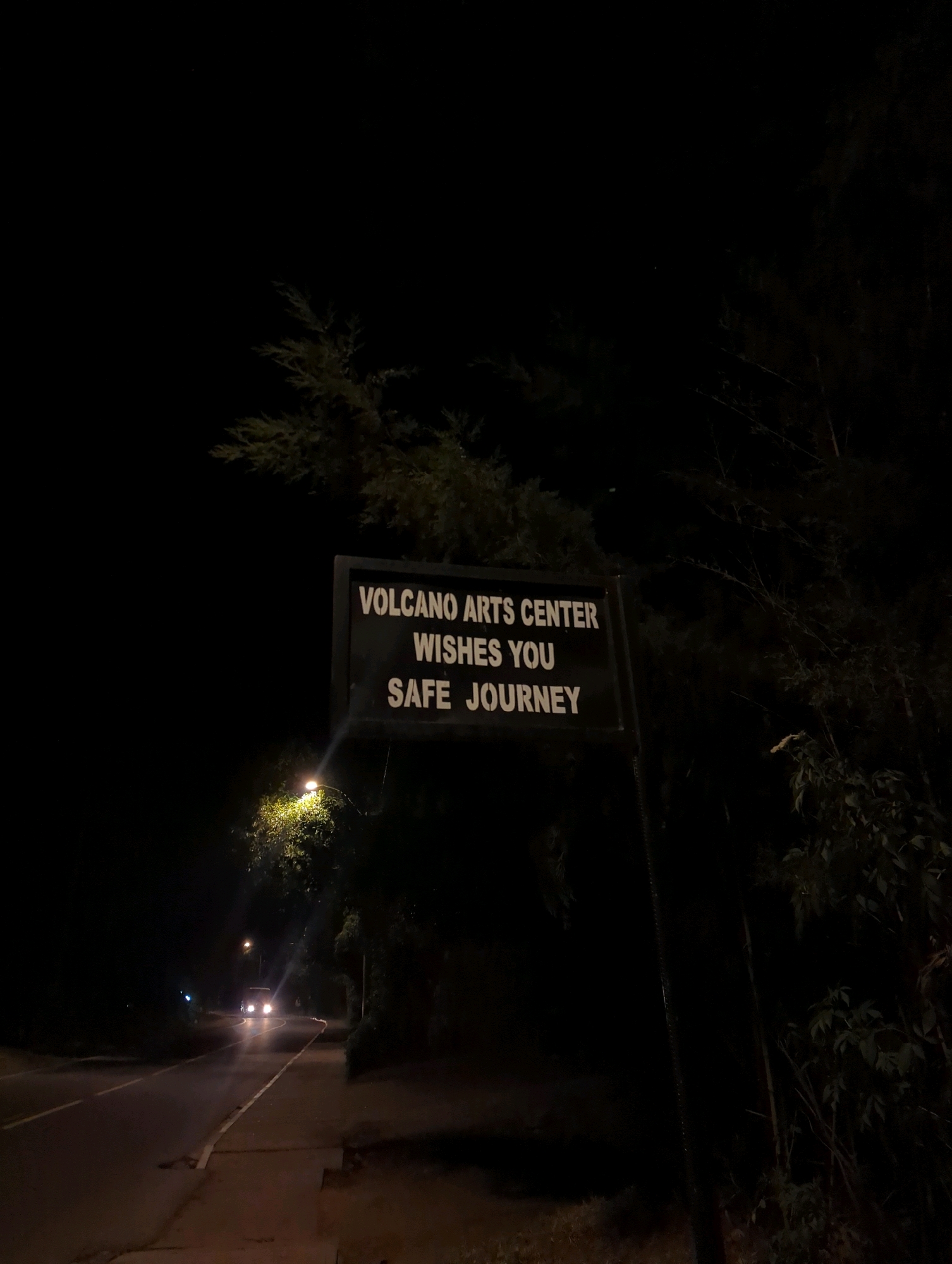 The image captures a nighttime scene on a road in Musanze District, Rwanda, near the Volcano Arts Center. A dark sign with white lettering reads "VOLCANO ARTS CENTER WISHES YOU SAFE JOURNEY". The sign is illuminated by a nearby streetlamp, casting a glow on the surrounding foliage. In the background, a vehicle with its headlights on travels along the asphalt road, which has a paved sidewalk on the right. Trees and dense vegetation line the road, creating a dark and somewhat mysterious atmosphere. The overall impression is one of a quiet, nocturnal setting in a semi-urban or rural area of Musanze.