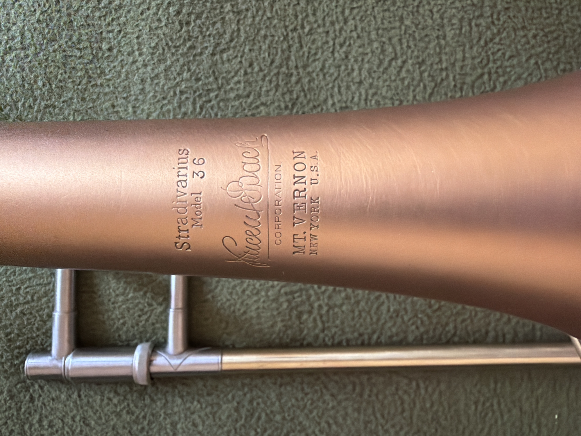 This close-up shot features the bell and a portion of the slide assembly of a brass instrument, specifically a trombone, resting on a textured, dark green fabric surface. No people are visible. The instrument's bell appears to be made of rose brass or a similar copper-toned alloy, exhibiting a brushed finish with some faint surface marks. The slide components, including the tuning slide and brace, are silver-colored metal.
Prominently engraved vertically on the bell are several lines of text. The largest text reads "Stradivarius" and "Model 36". Below this is the stylized signature "Vincent Bach", followed by "CORPORATION.". Further down, in smaller capital letters, is "MT. VERNON" and "NEW YORK U.S.A.". The instrument is stationary, with no activity taking place. The background is softly out of focus, emphasizing the details of the trombone. While the image is provided with a location context of Kyiv, Ukraine, the visible text indicates the instrument's manufacturing origin in New York, U.S.A. The time of day cannot be determined from the lighting.