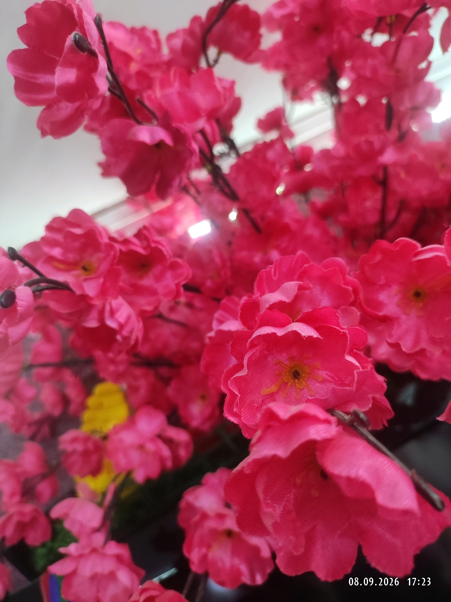 The image shows a close-up view of artificial pink flowers. The flowers are arranged on dark branches, and some yellow artificial flowers are visible in the background. The lighting suggests an indoor setting, possibly a shop or a decorative display. The bottom right corner of the image contains a timestamp: "08.09.2026 17:23". There is no information in the image to suggest a location in Makhachkala, Russia.