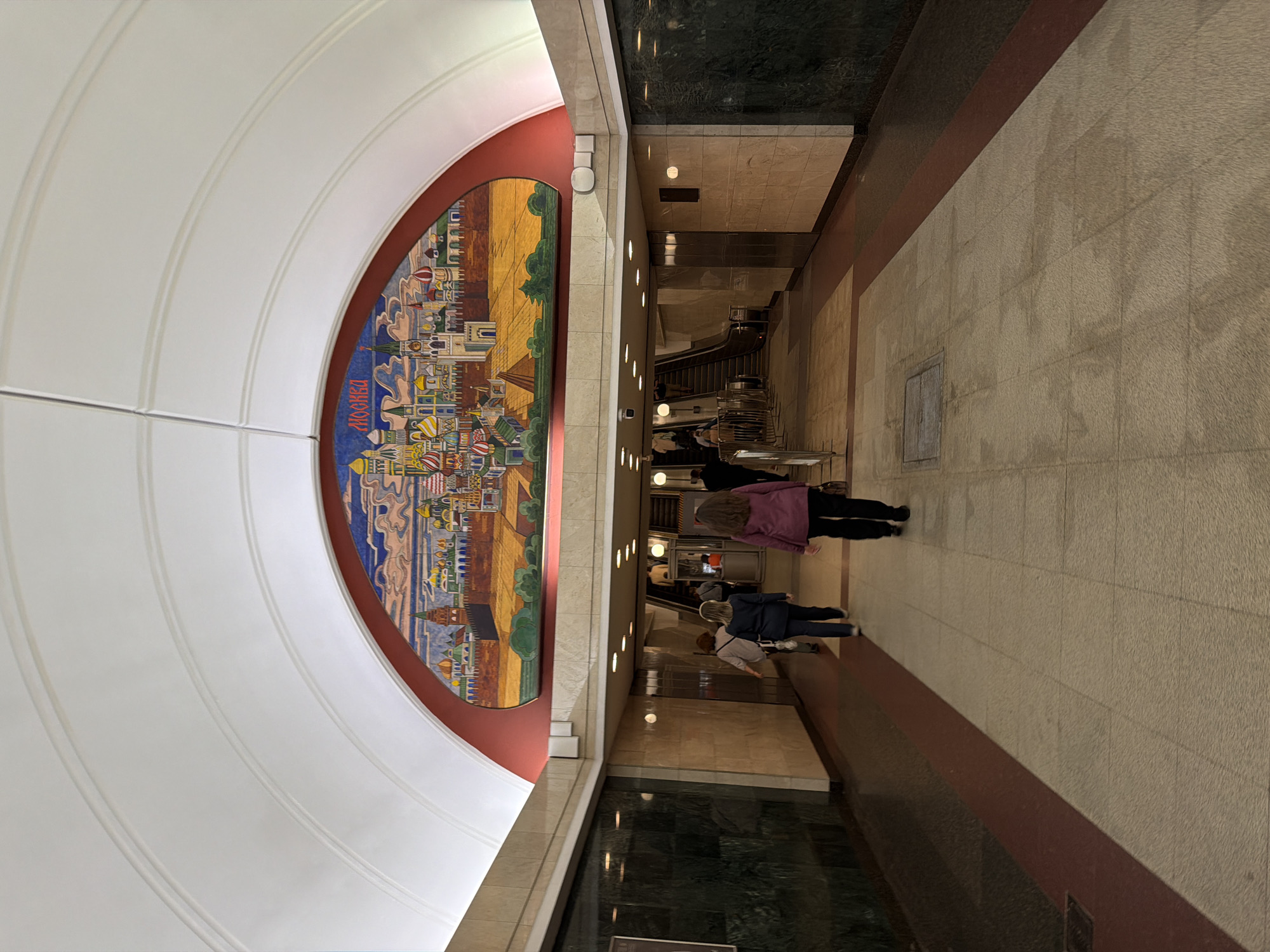 The image depicts the interior of a Moscow Metro station, likely at the Prospekt Mira station, which is known for its mosaics. The focal point of the scene is a large, semicircular mosaic adorning the station's vaulted ceiling, depicting a colorful panorama of Moscow's historic cityscape, including numerous onion-domed churches and Kremlin-like structures. The word "МОСКВА" (Moscow) is visible within the mosaic.
In the foreground, the tiled floor of the station corridor is visible, with a darker red border along the edges. Several individuals are walking away from the camera, heading towards a set of escalators in the background. These individuals appear to be passengers using the metro. One woman in the foreground is wearing a pink jacket and dark pants. To her left, another woman is wearing a dark blue jacket and jeans. Further ahead, more people are visible ascending the escalators. The station walls are paneled with marble or granite, and recessed lighting illuminates the space. The overall atmosphere is that of a busy, functional public transportation hub with artistic embellishments.