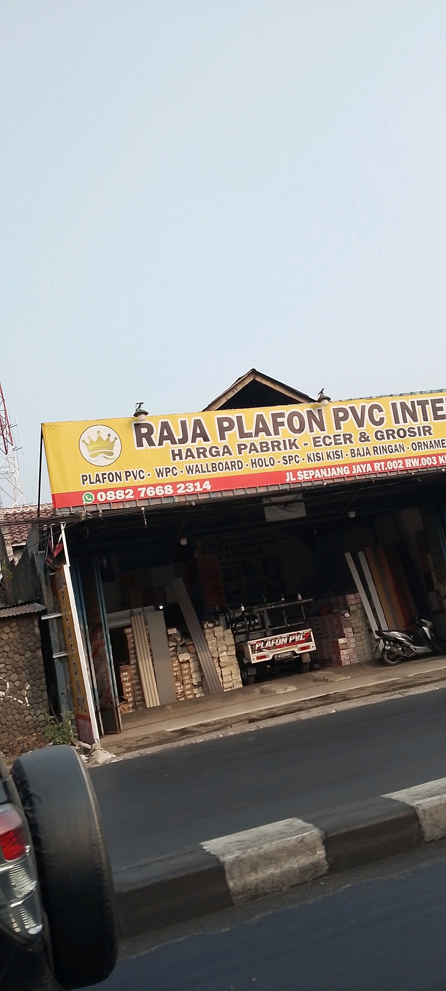 The image shows a storefront with a yellow banner that reads "RAJA PLAFON PVC INTERIOR". Below this, it lists "HARGA PABRIK - ECER & GROSIR" and "PLAFON PVC - WPC - WALLBOARD - HOLO - SPC - KISI KISI - BAJA RINGAN - ORNAMEN". A phone number "0882 7668 2314" and an address "JL SEPANJANG JAYA RT.002 RW.003 KOTA BEKASI" are also visible on the banner. The storefront appears to be a business selling building materials, specifically for ceilings and interiors, located in the Sepanjang Jaya area of Bekasi, Indonesia. The area appears to be a commercial street with a road and sidewalk in the foreground, and a parked motorcycle and a small truck are visible in front of the store. The sky is clear and bright.