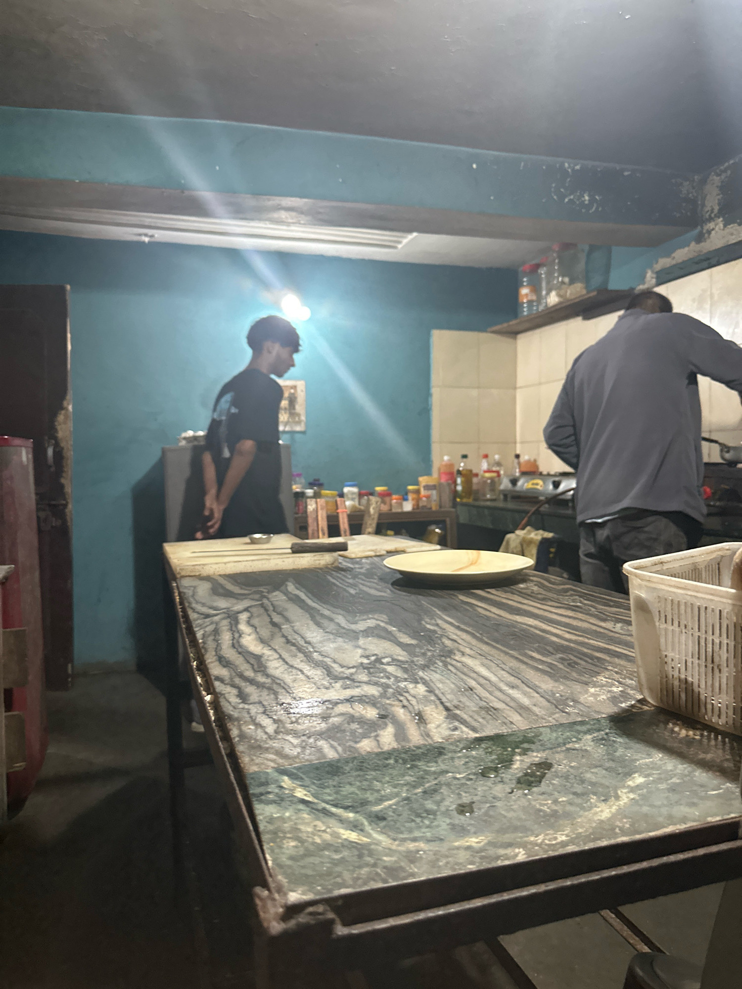In a dimly lit indoor space, possibly a kitchen or food preparation area, two individuals are visible. One person, wearing a dark t-shirt with a graphic on the back, stands with their hands clasped behind their back, facing away from the camera towards a turquoise wall. This individual is positioned near a counter. The second person, wearing a dark jacket and jeans, is seen from the back, engaged in cooking at a stove located on a counter against a tiled wall. The area is cluttered with various items, including bottles on shelves, containers, and cooking utensils. A prominent marble-topped counter with a dark, patterned surface occupies the foreground, with a plate on it. The overall environment appears utilitarian and somewhat aged, with visible wear on the walls and ceiling.