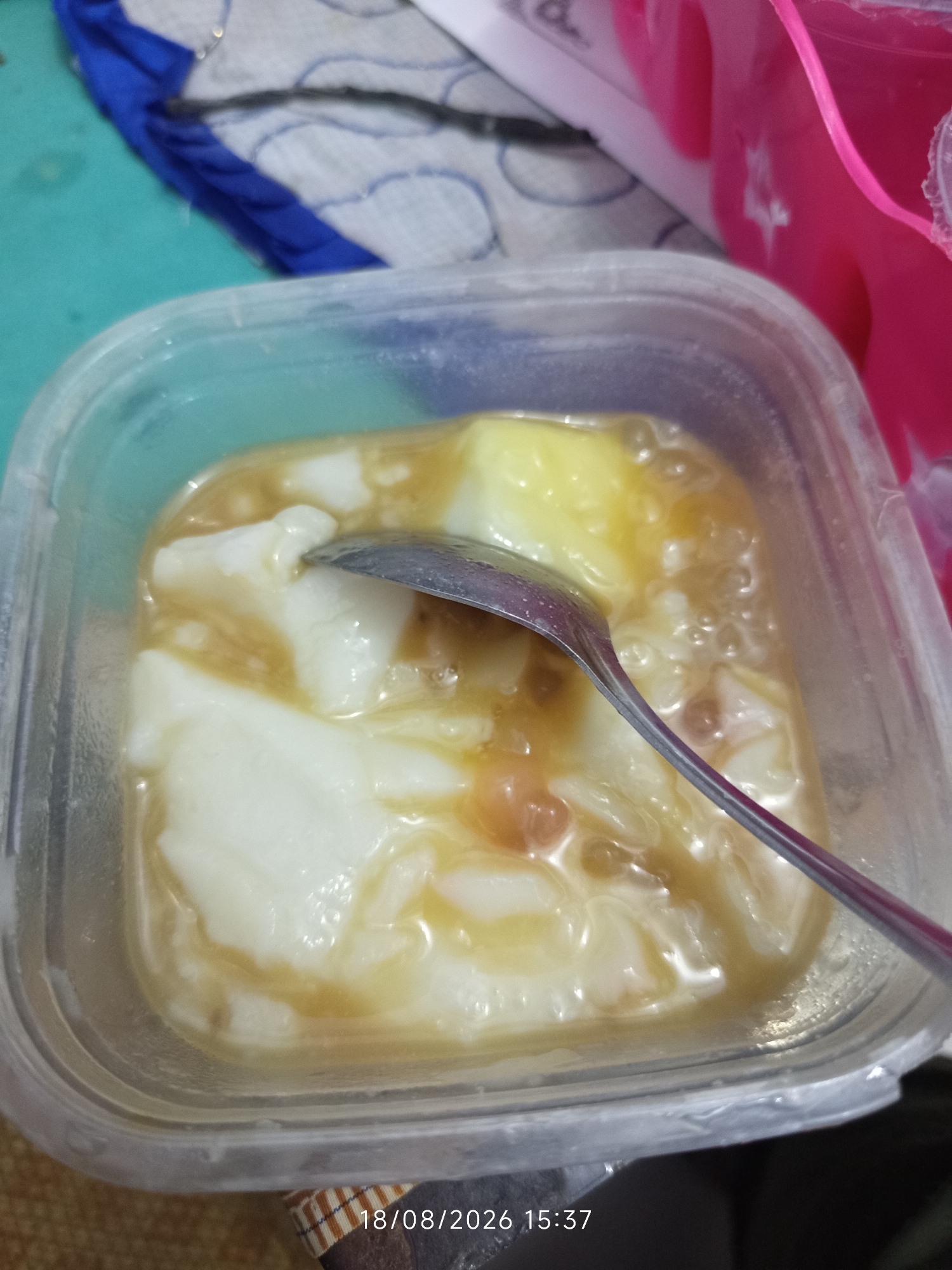 A close-up, overhead shot shows a clear plastic container filled with a dessert. The dessert appears to be a creamy, white base, possibly tofu pudding or a similar sweet dish, covered in a light brown, syrupy sauce. A metal spoon is partially submerged in the dessert, suggesting it is being eaten or has recently been consumed. The container is slightly fogged on the inside, indicating it may have been refrigerated. The background is blurred, but hints of teal and pink objects, along with fabric textures, are visible. A timestamp at the bottom of the image reads "18/08/2026 15:37". The image appears to have been taken in a domestic setting, possibly a kitchen or dining area. There are no people visible in the frame.