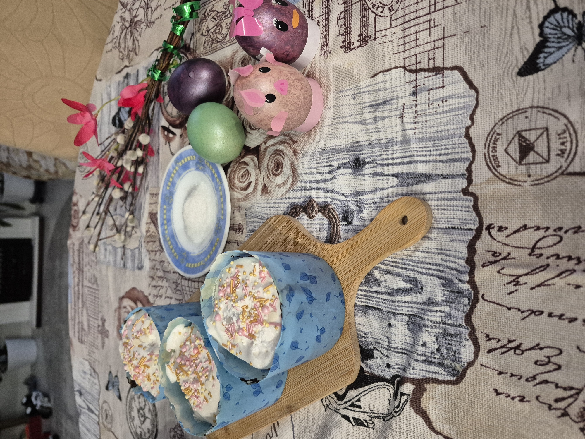 The image shows a table setting, likely for an Easter celebration. On a tablecloth with a print resembling old maps and script, there is a wooden cutting board with three Easter cakes (kulich) wrapped in light blue paper and decorated with white icing and colorful sprinkles. Next to the cutting board, there is a small plate. Near the top of the image are several Easter eggs, some of which are painted to resemble animals (pig and bird). There is also a small bouquet of willow branches and pink flowers. The location is likely Chaikovsky, Russia, based on the user's query. The setting appears to be indoors, possibly a dining room or kitchen.
