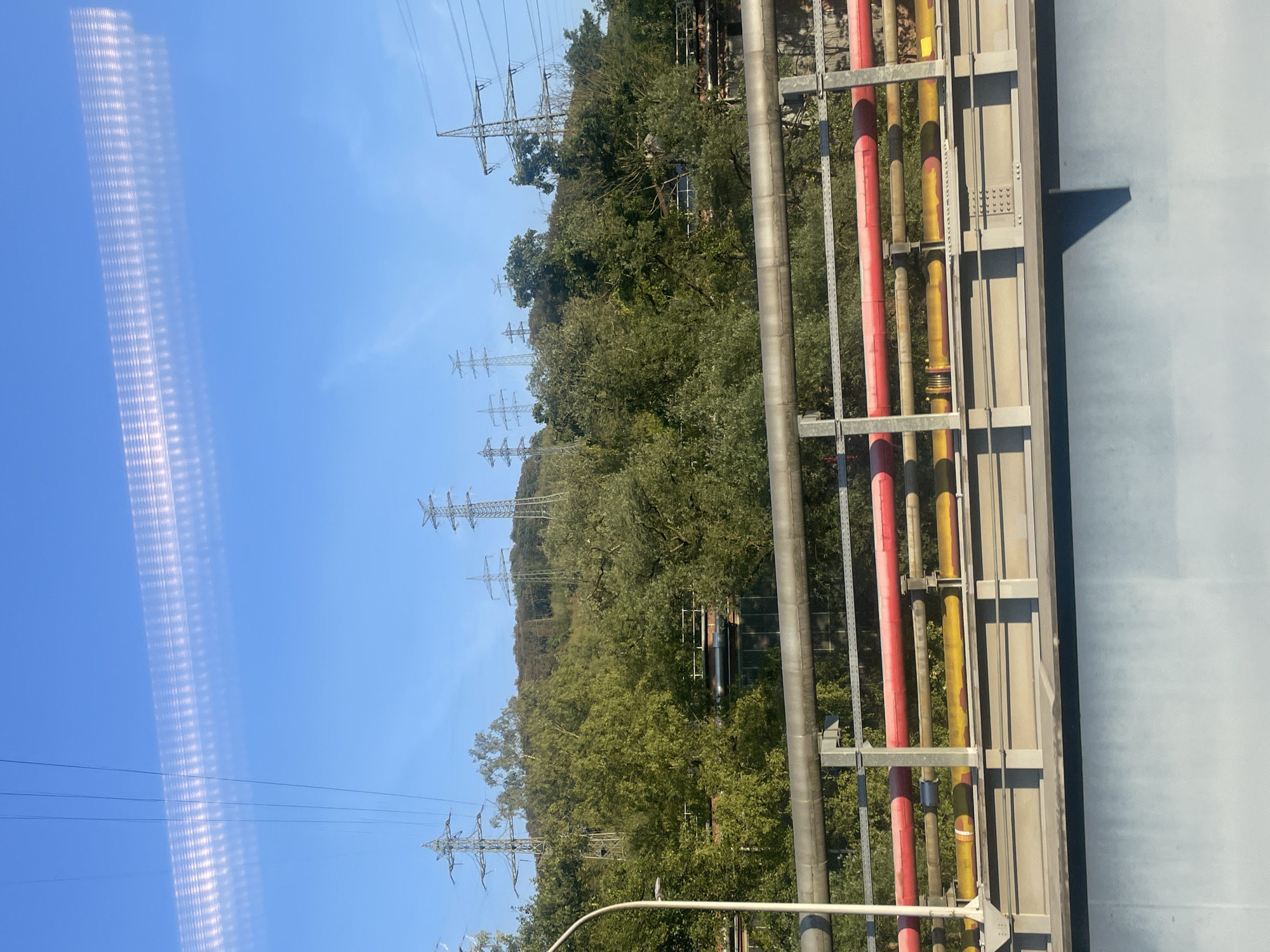 The image depicts an industrial landscape with electrical pylons and pipelines set against a backdrop of lush green trees and a clear blue sky. The foreground features a bridge with several large pipes, some painted red and others yellow, supported by metal structures. A vertical pole, possibly a lamppost, is visible on the left side of the bridge. The middle ground is dominated by dense foliage, suggesting a wooded area, with the industrial structures and pylons rising above the trees. In the background, multiple electricity pylons of varying sizes are scattered across a hillside. The sky above is bright and clear, with a distinct, horizontal band of reflected light that appears as a textured, luminous strip across the top of the image, likely a reflection from the camera lens or window. The scene is devoid of human presence. This view is indicative of the industrial areas surrounding the city of Iserlohn, Germany, which is known for its hilly terrain and proximity to industrial facilities, often featuring such infrastructure.