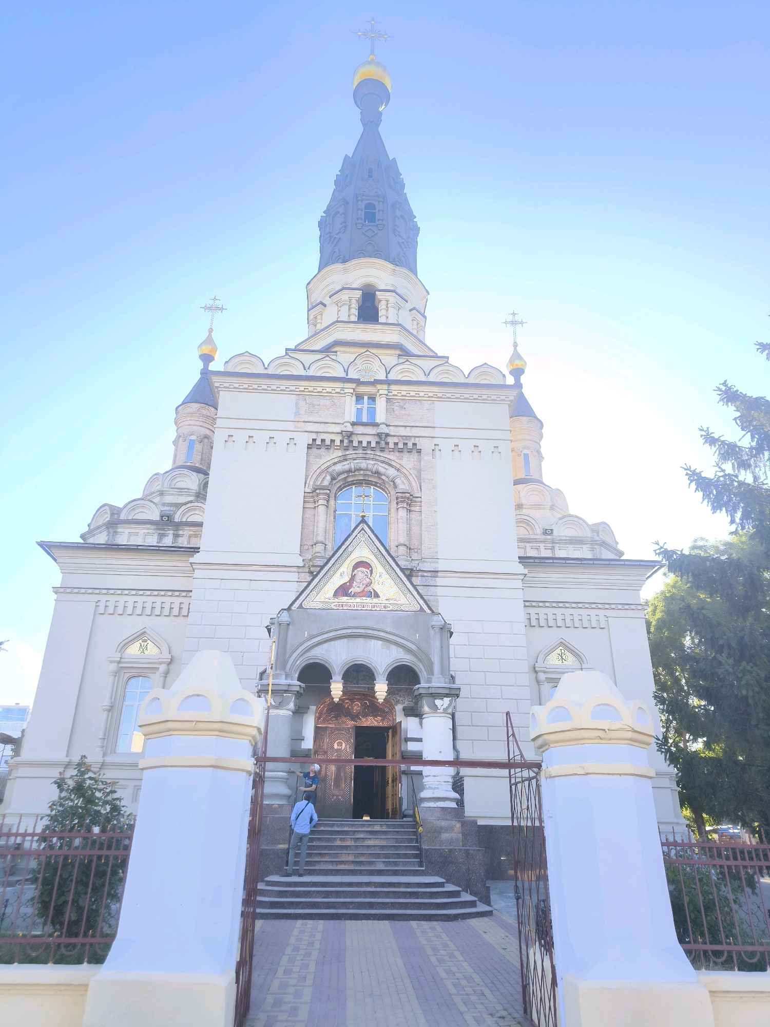 An exterior, low-angle, full shot captures the front of a grand, light-colored Orthodox church under a clear blue sky. The church features multiple tiered domes topped with golden crosses, a tall central spire, and intricate architectural details including arched windows and decorative stonework. A mosaic icon is visible above the main entrance, which is recessed and features ornate doors. Steps lead up to the entrance, and a person in a blue shirt and grey pants is ascending them, facing away from the camera, holding a dark bag. Another person in a dark shirt and hat is standing on the stairs. Two decorative white pillars with yellow accents flank a metal fence in the foreground, partially obscuring the view of the church entrance and steps. Trees are visible to the right of the church. The church is identified as St. Nicholas Cathedral in Mykolaiv, Ukraine.