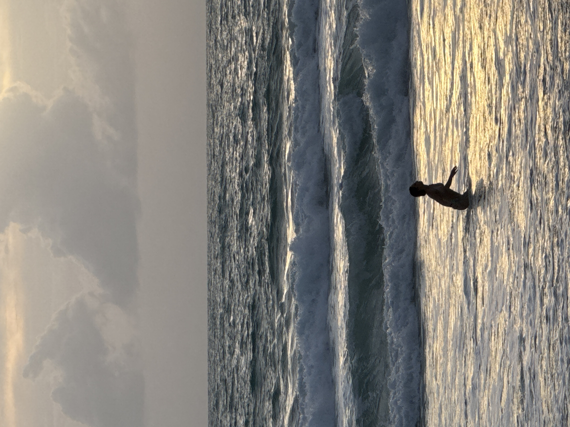 A lone person, silhouetted against the golden light reflecting on the water, stands in the ocean at the coast of Choeng Thale, Thailand. The person is facing left, with their head tilted upwards, appearing to be looking at the sky. Waves are breaking around them, with white foam cresting on the dark water. The sky above is overcast with diffused light filtering through the clouds, suggesting either sunrise or sunset. The overall scene is one of natural beauty and solitude.
