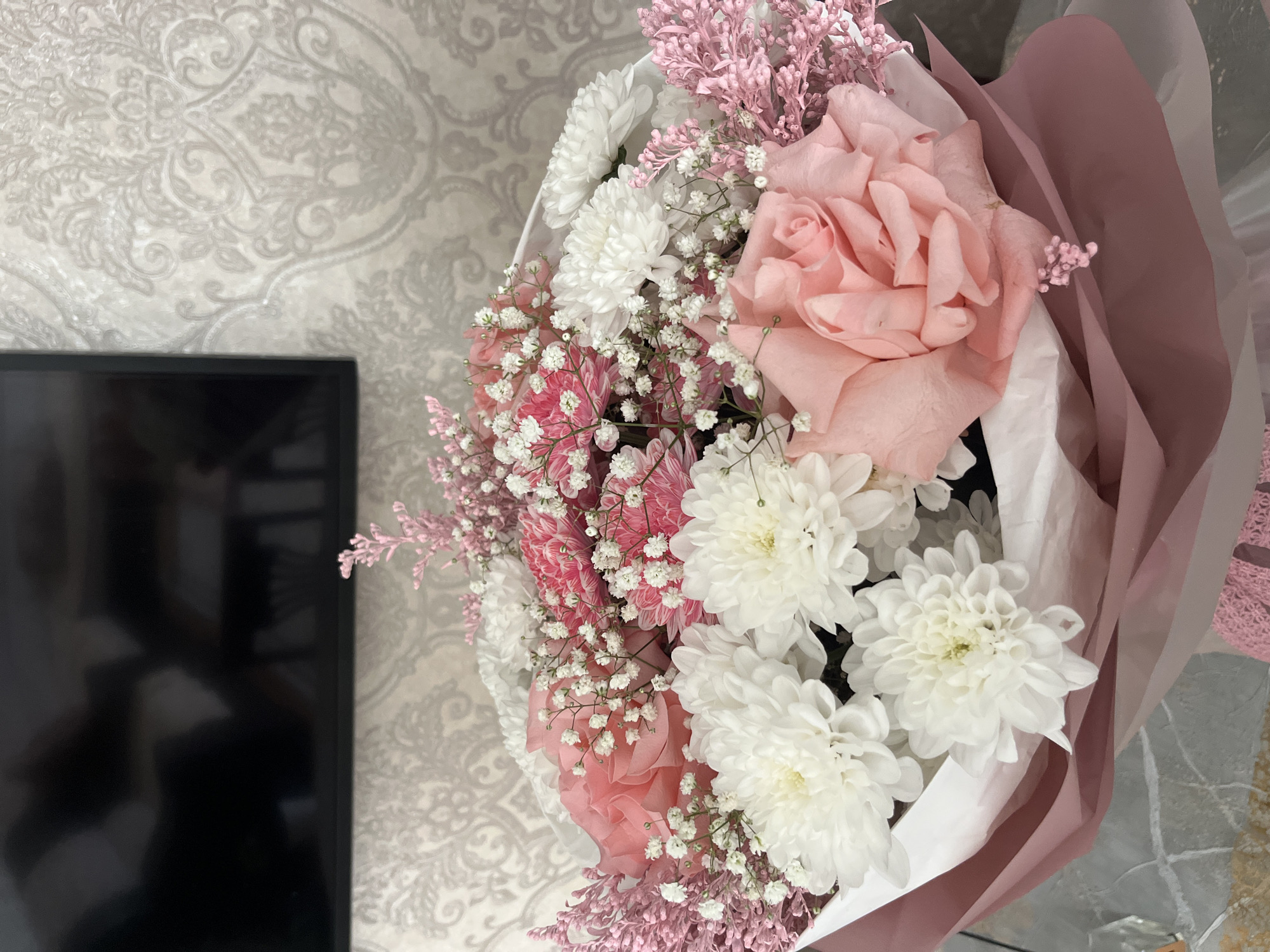 The image is a close-up of a bouquet of flowers. The bouquet contains pink roses, white chrysanthemums, and pink and white baby's breath. The flowers are wrapped in pink and white paper. The background includes a patterned wall and a black television screen. There is no evidence to suggest the location is Kaspiysk, Russia. No people, interactions or actions are visible in the image.