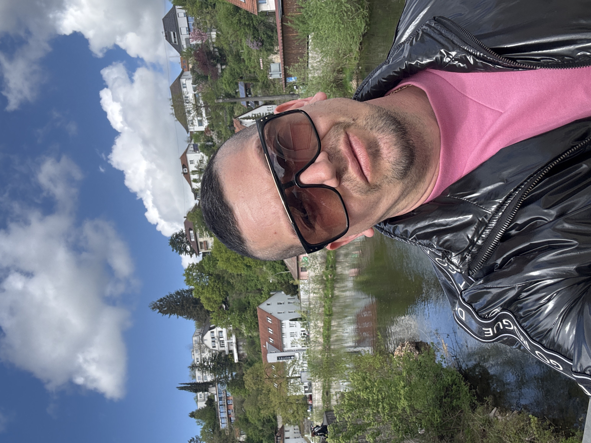 The image shows a man wearing sunglasses, a pink shirt, and a shiny black jacket with the word "GUESS" on the sleeve. He appears to be taking a selfie with a bridge in the background. The location seems to be Backnang, Germany, identifiable by the buildings and water in the background which match its urban landscape. There is vegetation near the river or canal, and the sky shows blue with white clouds. The man's face is partially visible behind the sunglasses.