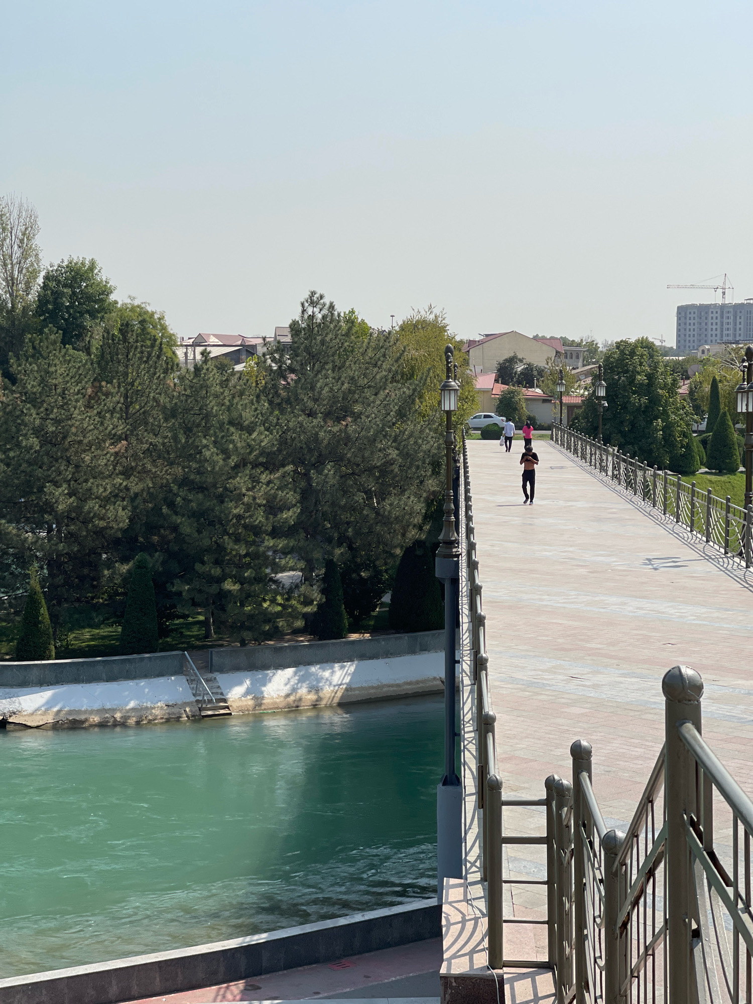 This image shows a pedestrian bridge over a canal in Tashkent, Uzbekistan. The bridge is made of concrete and has metal railings. There are several people on the bridge, walking and jogging. The canal is filled with green water. In the background, there are trees and buildings. The sky is clear and blue. The overall scene depicts a pleasant day in a city park.