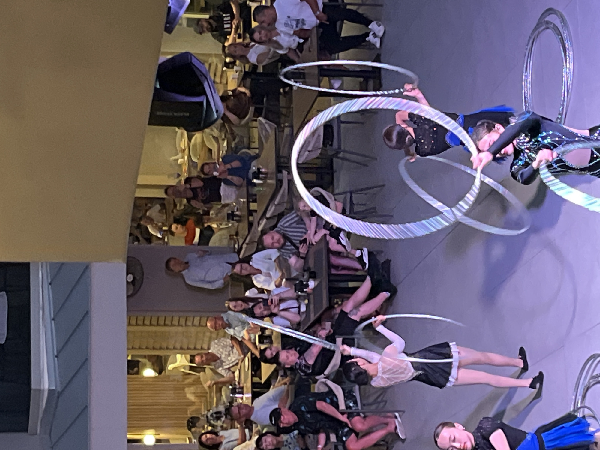 The image depicts a performance involving two women manipulating hoops, possibly in a show or entertainment setting. The audience, comprised of diverse individuals sitting at tables and watching the performance. The location appears to be a dining area, potentially in a restaurant. Based on external information, it is likely that the location is in the city of Alanya, Turkey.