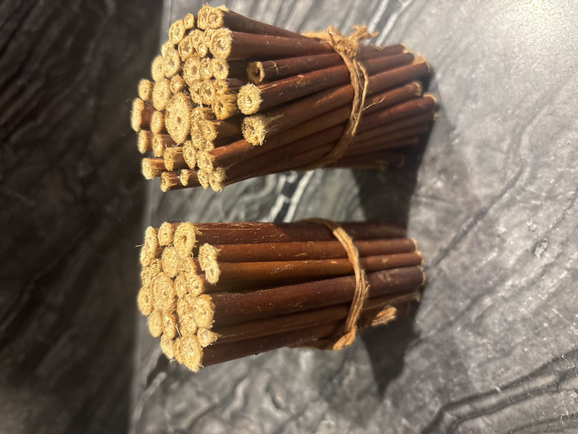 Two bundles of dried twigs, each tied with twine, are placed on a mottled gray countertop. The twigs are a reddish-brown color and have been cut to reveal hollow, beige interiors. The background is a dark, textured surface that appears to be a wall or backsplash. The lighting creates shadows that accentuate the texture of the countertop and the twigs. The scene appears to be indoors, and there is no indication of the city of Dubai or the United Arab Emirates.