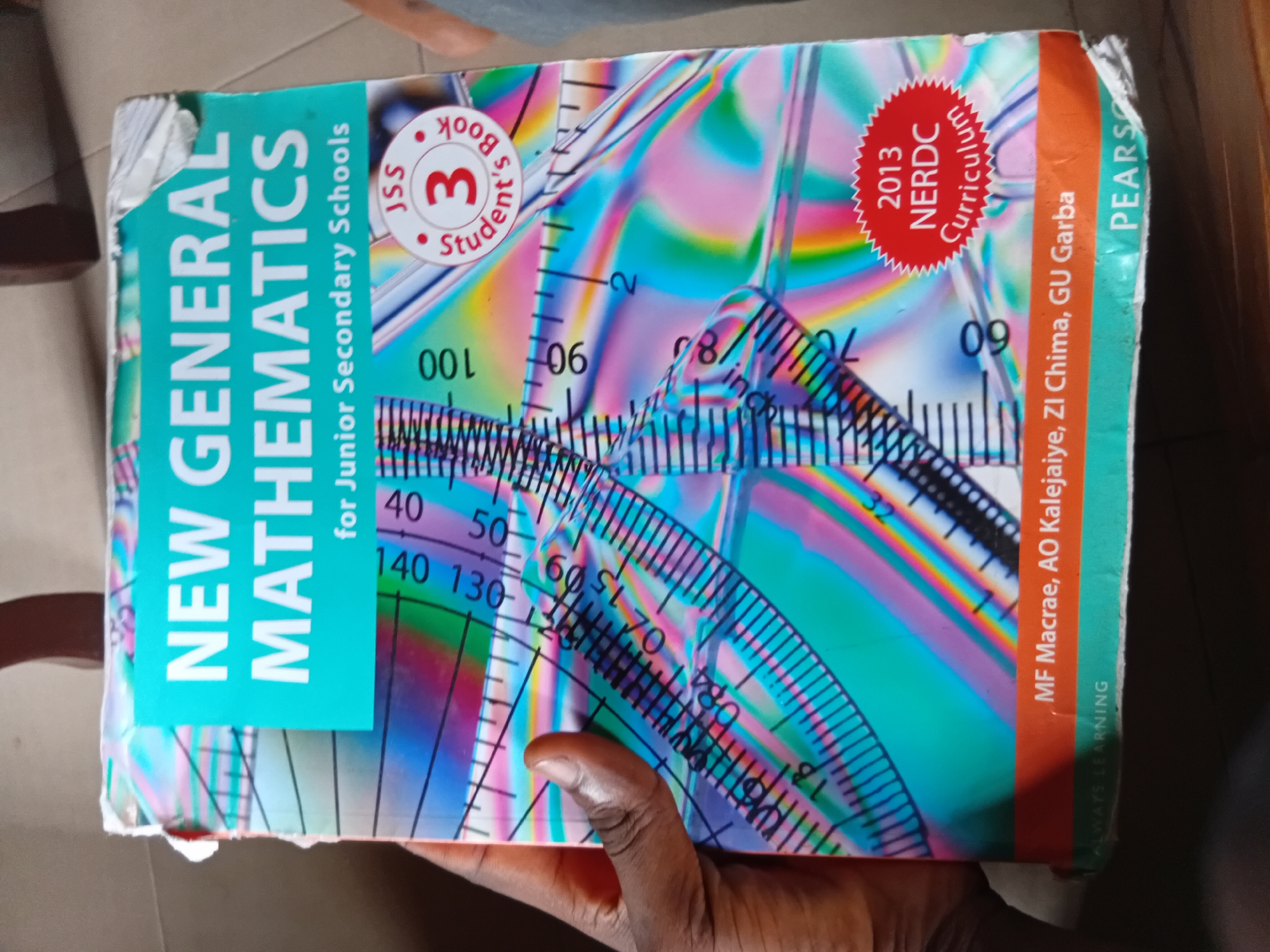 The image shows the cover of a mathematics textbook. The title, "NEW GENERAL MATHEMATICS for Junior Secondary Schools," is prominently displayed. The cover features a colorful, abstract background with geometric patterns and rulers, suggesting the subject matter. A hand is visible on the left side of the image, holding the book. In the bottom right corner, there is a circular red badge with "2013 NERDC Curriculum" written on it, indicating the book's publication year and curriculum. Below the title, the authors' names are listed as MF Macrae, AO Kalejaiye, ZI Chima, and GU Garba. At the very bottom, the publisher's name, "PEARSON," is visible. The book appears to be in a private setting, possibly a home or office, as indicated by the blurred background and what looks like furniture. The environment is indoors and appears to be lit by artificial light or natural light coming from an unseen source. The overall scene is focused on the textbook itself.