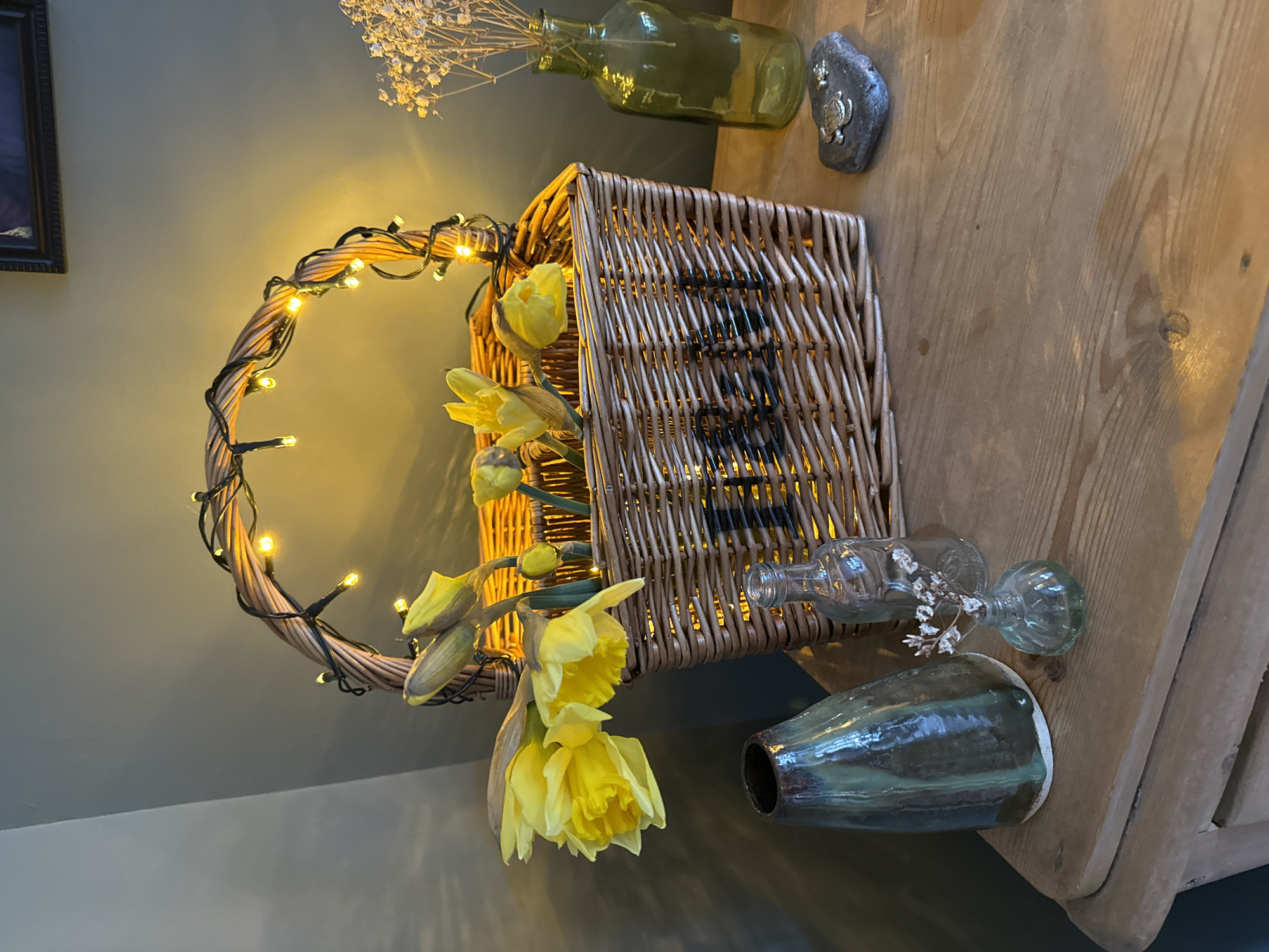 The image depicts a still life arrangement. A woven basket, adorned with fairy lights, contains and is surrounded by bright yellow daffodils. Next to the basket and slightly behind it, a glass vase holds dried baby's breath flowers. A decorative stone sits nearby. In the foreground, a small glass bottle and a larger ceramic vase are placed. The scene is set on a wooden surface, likely a table, against a muted green wall. The photograph seems to be taken indoors in East Suffolk, United Kingdom, with the light suggesting either morning or early evening.