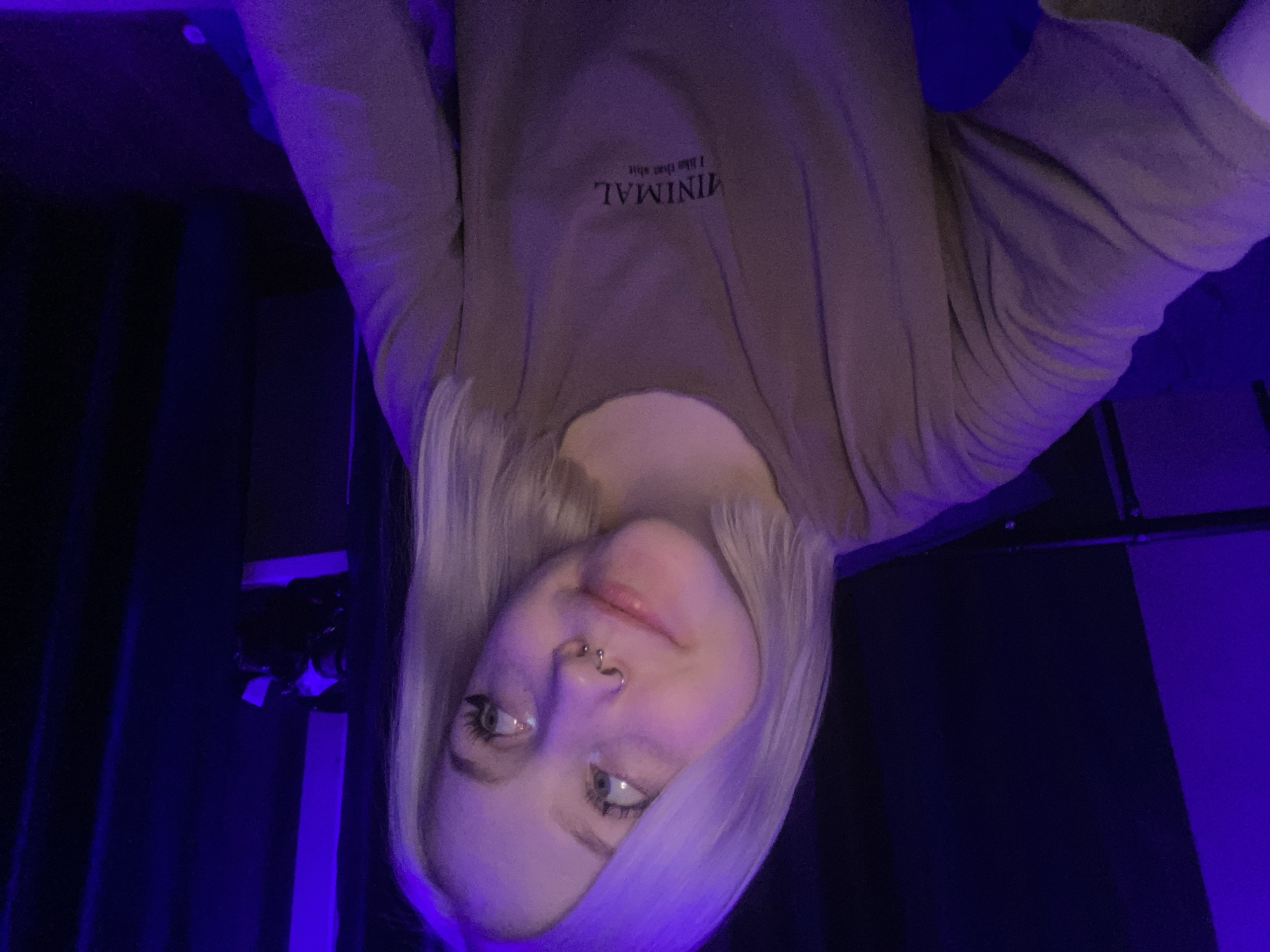 A young woman with long, platinum blonde hair poses for a close-up shot in a room illuminated with blue light. She wears a tan shirt with the word "MINIMAL" printed on it. She has fair skin and facial piercings in her nose. The background is dark, with hints of furniture and curtains. It is impossible to identify the location in the city of Izhevsk, Russia based solely on the provided image.