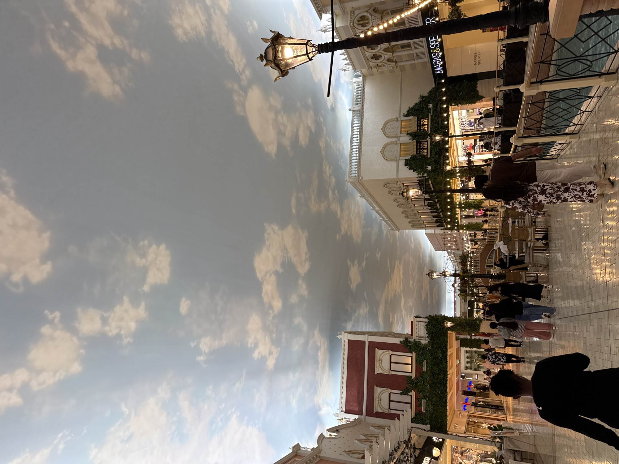The image depicts a scene at Villaggio Mall in Al Rayyan, Qatar. The architectural design mimics a Venetian street, with buildings lining a wide walkway under a painted sky-like ceiling. Several people are walking and interacting in the space. Some are standing near shops, while others are passing by. A lamppost is visible on the left, contributing to the faux-outdoor aesthetic. Shops and businesses are visible in the background, including Marks & Spencer.