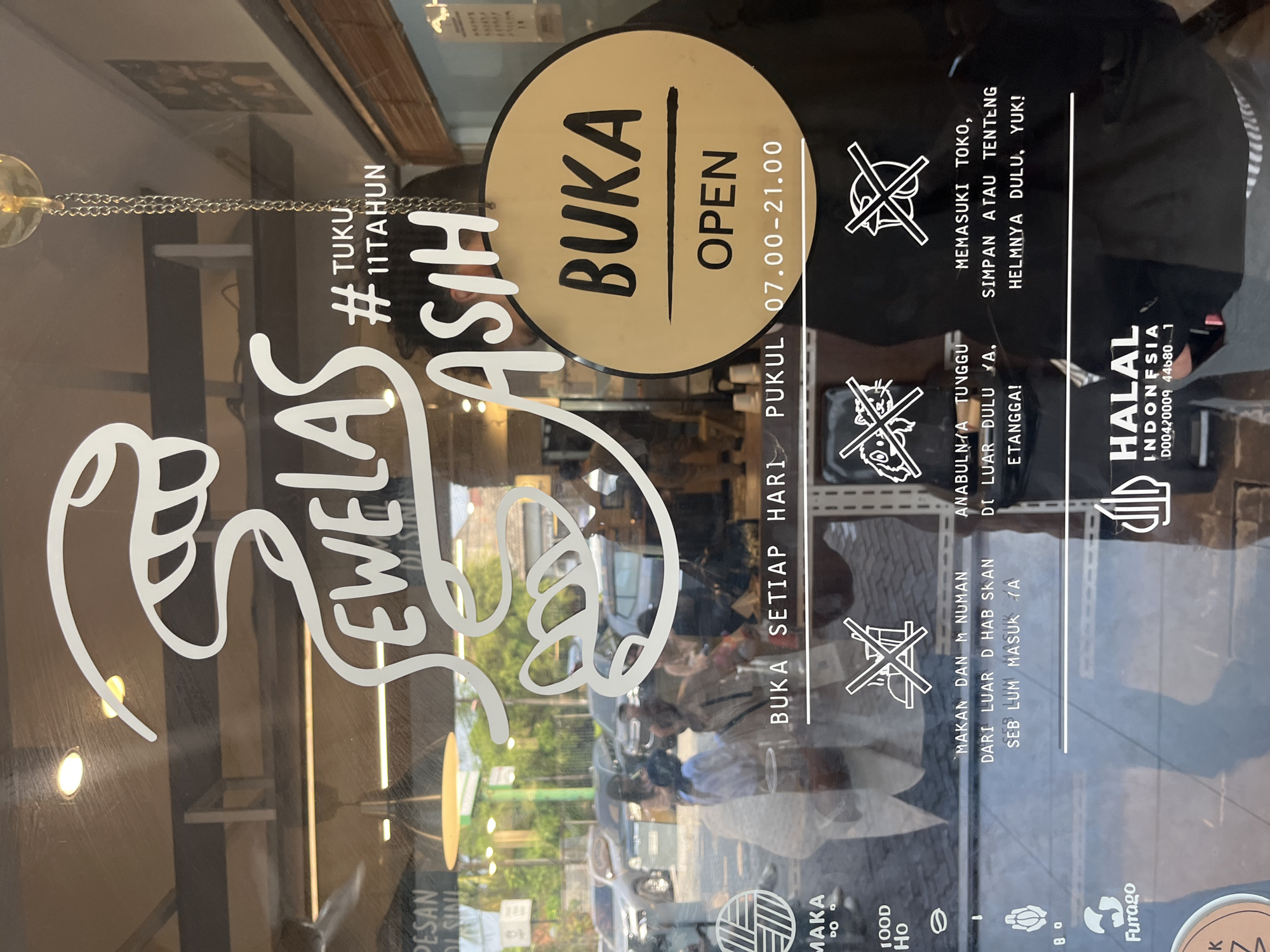 The image shows a glass storefront with white lettering and graphics. The prominent text reads "JEWELAS # TUKU 11 TAHUN ASIH". A round sign with the text "BUKA OPEN 07.00-21.00" is hanging on the glass. Below this, there are several graphic icons with accompanying text in Indonesian. One icon shows a crossed-out food item, with text "MAKAN DAN MINUMAN DARI LUAR DI HABISKAN SEBELUM MASUK YA". Another icon depicts crossed-out animals, and text "ANABULNYA TUNGGU DI LUAR DULU YA, ETANGGA!". A third icon shows a crossed-out bird, with text "MEMASUKI TOKO, SIMPAN ATAU TENTENG HELMNYA DULU, YUK!". The bottom of the storefront displays a "HALAL INDONESIA D00420009 44680_1" certification. The reflection on the glass reveals an outdoor scene with cars and people, suggesting a street-level establishment in a city. The overall impression is a business, likely a shop or restaurant, with clear operational information presented to customers.