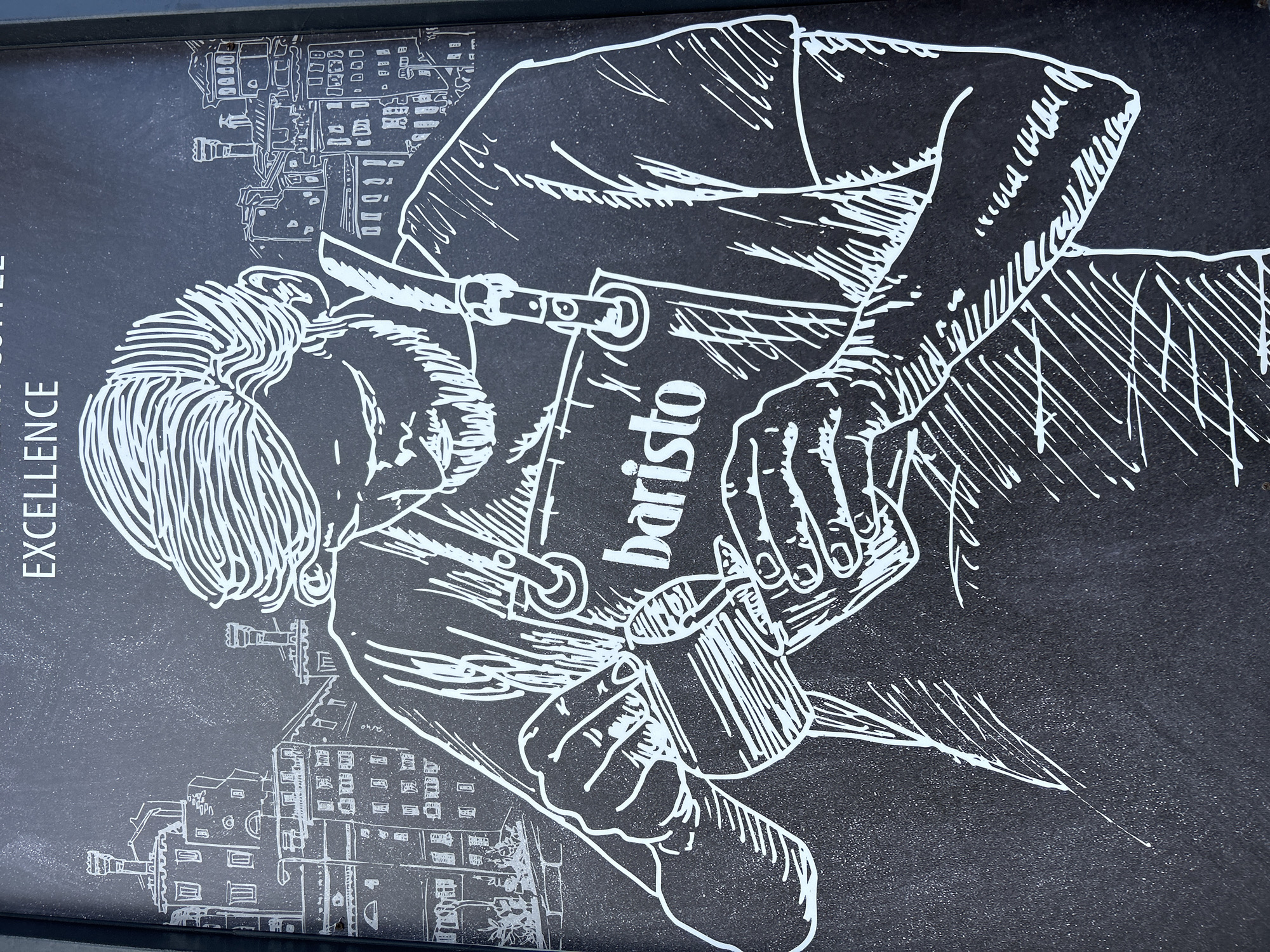 The image is a close-up of a sign, possibly a chalkboard or painted surface, featuring a white line drawing on a black background. The drawing depicts a barista preparing coffee. The barista is seen in profile, with their head and upper torso visible. They are wearing an apron that has the word "baristo" vertically printed on it. The barista is holding a cup in one hand and appears to be operating a coffee-making device (possibly a portafilter or milk frother) with the other. The drawing includes schematic representations of buildings, suggesting a cityscape in the background. The word "EXCELLENCE" is partially visible along the left edge of the image. The image appears to be located in Plovdiv, Bulgaria.