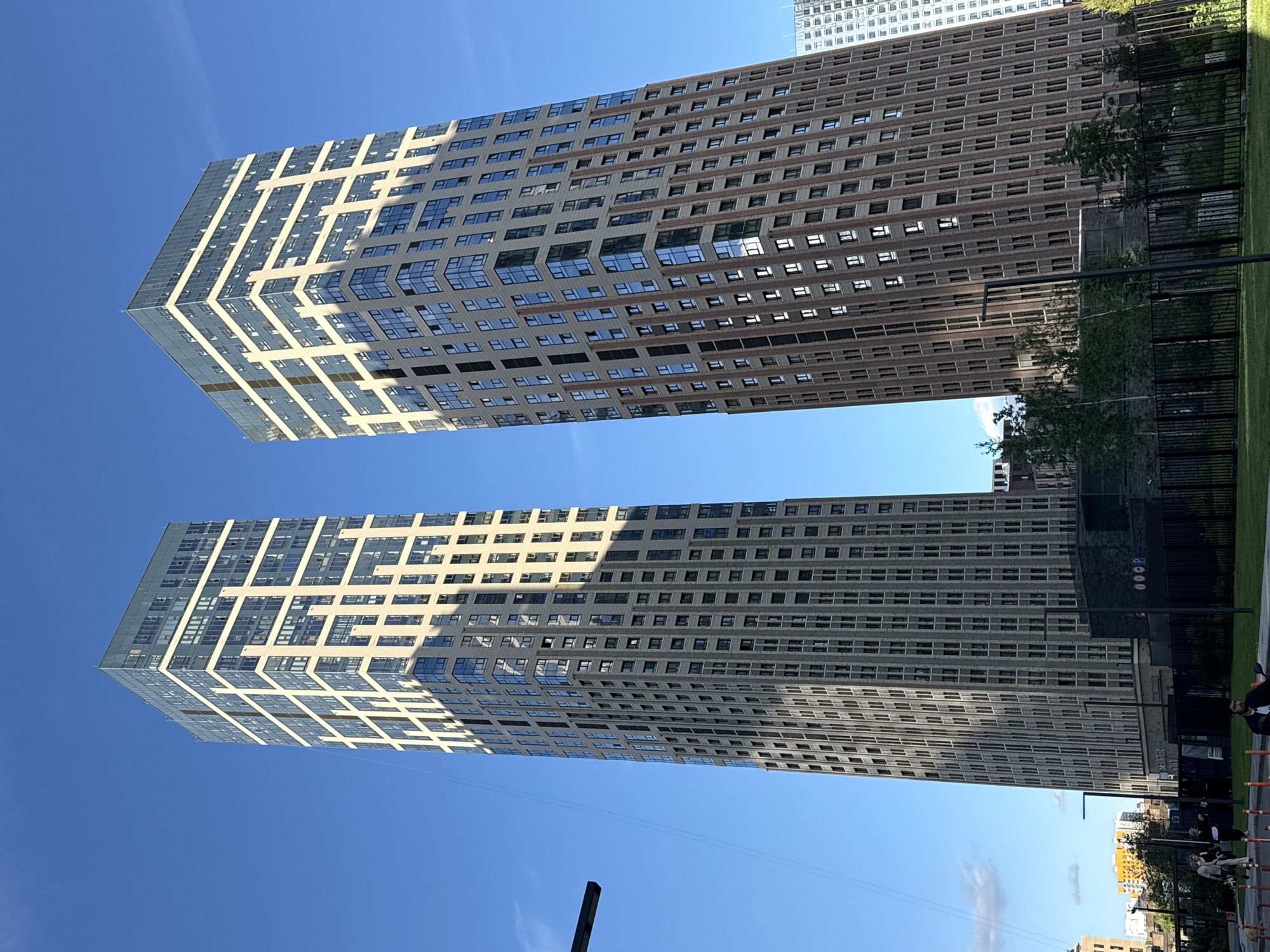 Two identical, modern skyscrapers stand tall against a clear blue sky. The buildings are characterized by their beige and dark brown facades, with numerous windows arranged in a grid pattern. The top of each skyscraper is capped with a glass structure. In the foreground, there is a black metal fence and some green grass. Several trees are also visible. A few people are walking in the lower left portion of the image, and one person is sitting on a scooter in the lower right. The scene appears to be a contemporary urban environment.
