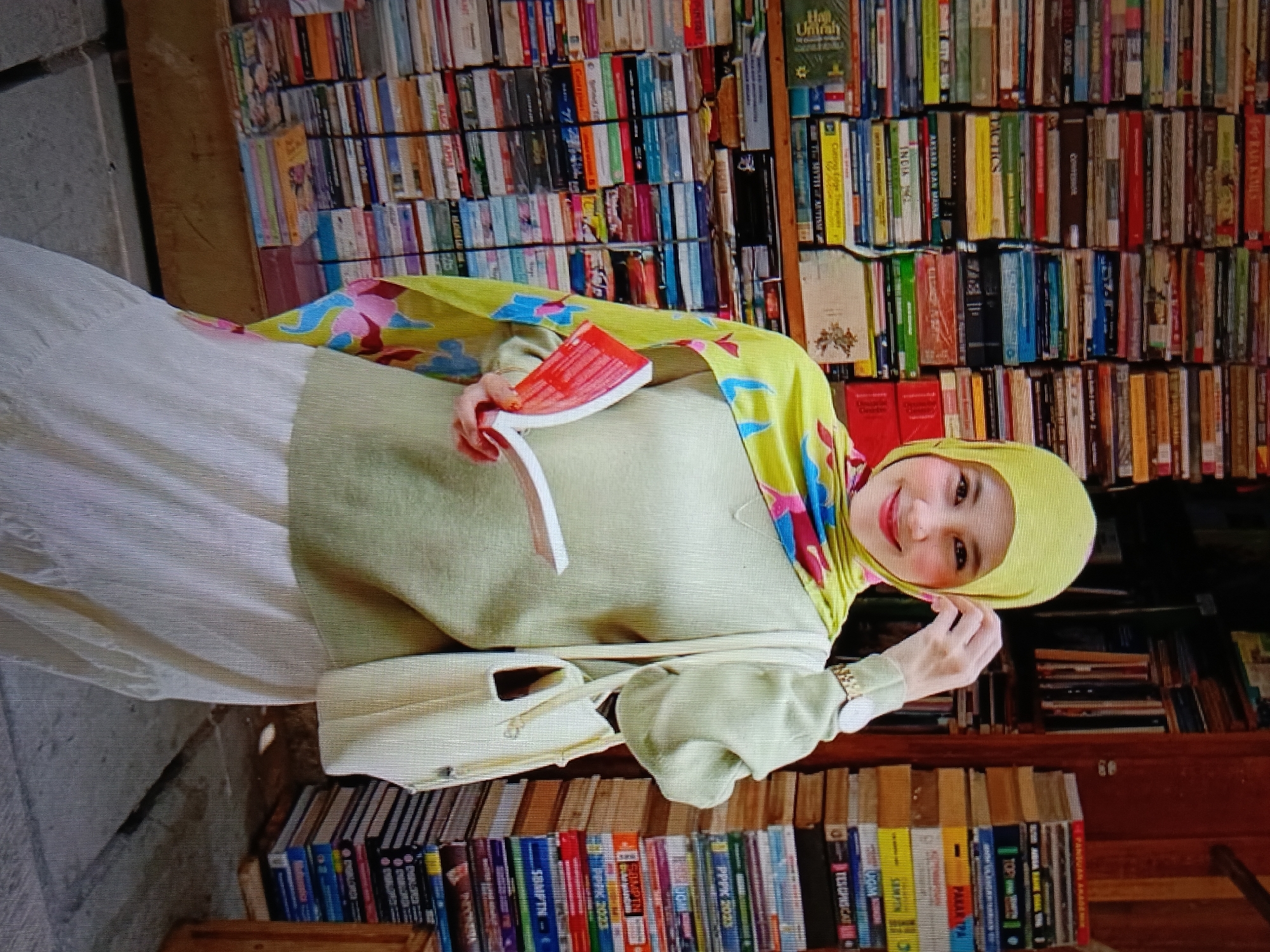 A woman wearing a yellow hijab and a light green outfit is seated in front of a bookshelf filled with books. She is holding a red book in her hands and looking at the camera with a smile. She has a light-colored handbag on her left side. The scene appears to be indoors, possibly in a home library or a bookstore in Raha, Indonesia. The lighting is warm and natural, highlighting the textures of the books and the woman's clothing.