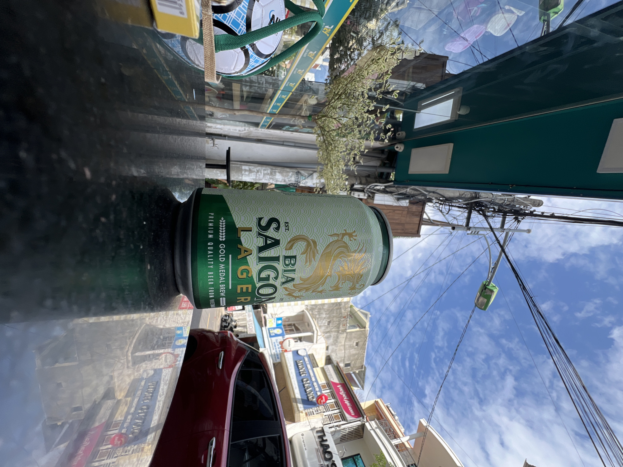 The image depicts an outdoor scene in Nha Trang, Vietnam, likely on a street or sidewalk with shops and buildings. In the foreground, a metallic, black surface, possibly a table, reflects the surroundings. A partially visible can of "BIA SAIGON LAGER" is prominent, its green and gold design featuring a dragon motif. To the left, a white and blue soccer ball in a woven basket with green handles is also visible. In the background, a red car is parked on the street. The architecture suggests a commercial area with shopfronts displaying signage, some in Vietnamese. Overhead, a network of electrical wires and a streetlamp are visible against a bright blue sky with scattered clouds. The presence of a streetlamp and the general urban environment indicate it's a public space within the city.