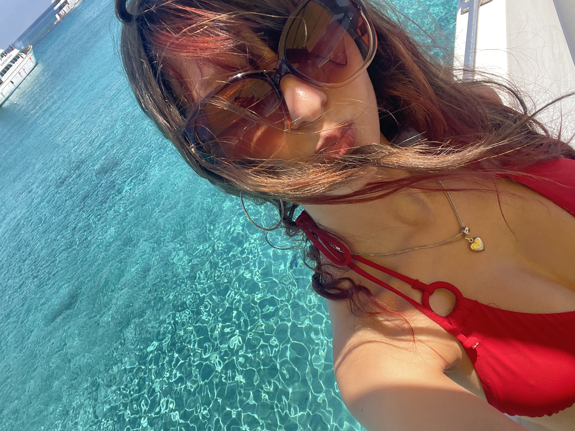 A woman is posing for a selfie in bright sunlight on a boat in the turquoise waters off the coast of Neo Chorio, Cyprus. She is wearing a red bikini top and large sunglasses, with her hair falling around her face. A silver necklace with a heart-shaped pendant is visible. The clear water reflects the sunlight, creating a shimmering effect. In the background, white boats are anchored in the distance. The scene suggests a leisurely day at sea.