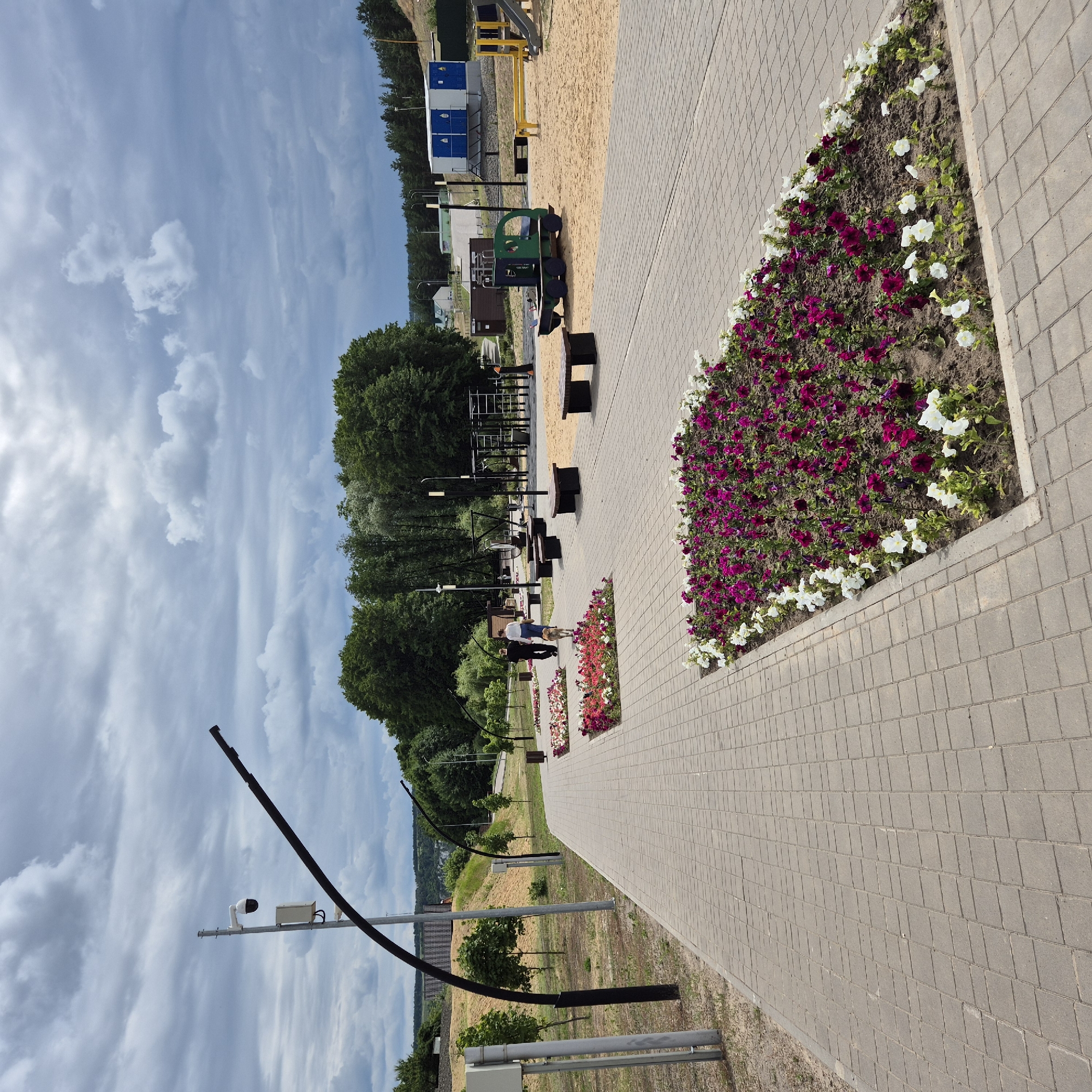 In the city of Liski, Russia, a wide, paved walkway extends into the distance, lined with manicured flower beds bursting with vibrant purple and white blossoms. The walkway is bordered by small, newly planted trees on one side and a sandy area on the other, which features a children's playground with swings, slides, and a toy train. A modern outdoor exercise area is also visible, equipped with various metal bars and equipment. Streetlights with curved black poles illuminate the path. In the background, lush green trees form a dense forest, and beyond that, buildings and residential structures are scattered across the landscape, hinting at the edge of the city. The sky is overcast with dramatic, dark clouds, suggesting an impending storm. A security camera is mounted on a pole near the walkway. Two individuals, a man and a woman, walk along the path accompanied by a dog, their figures receding into the distance.