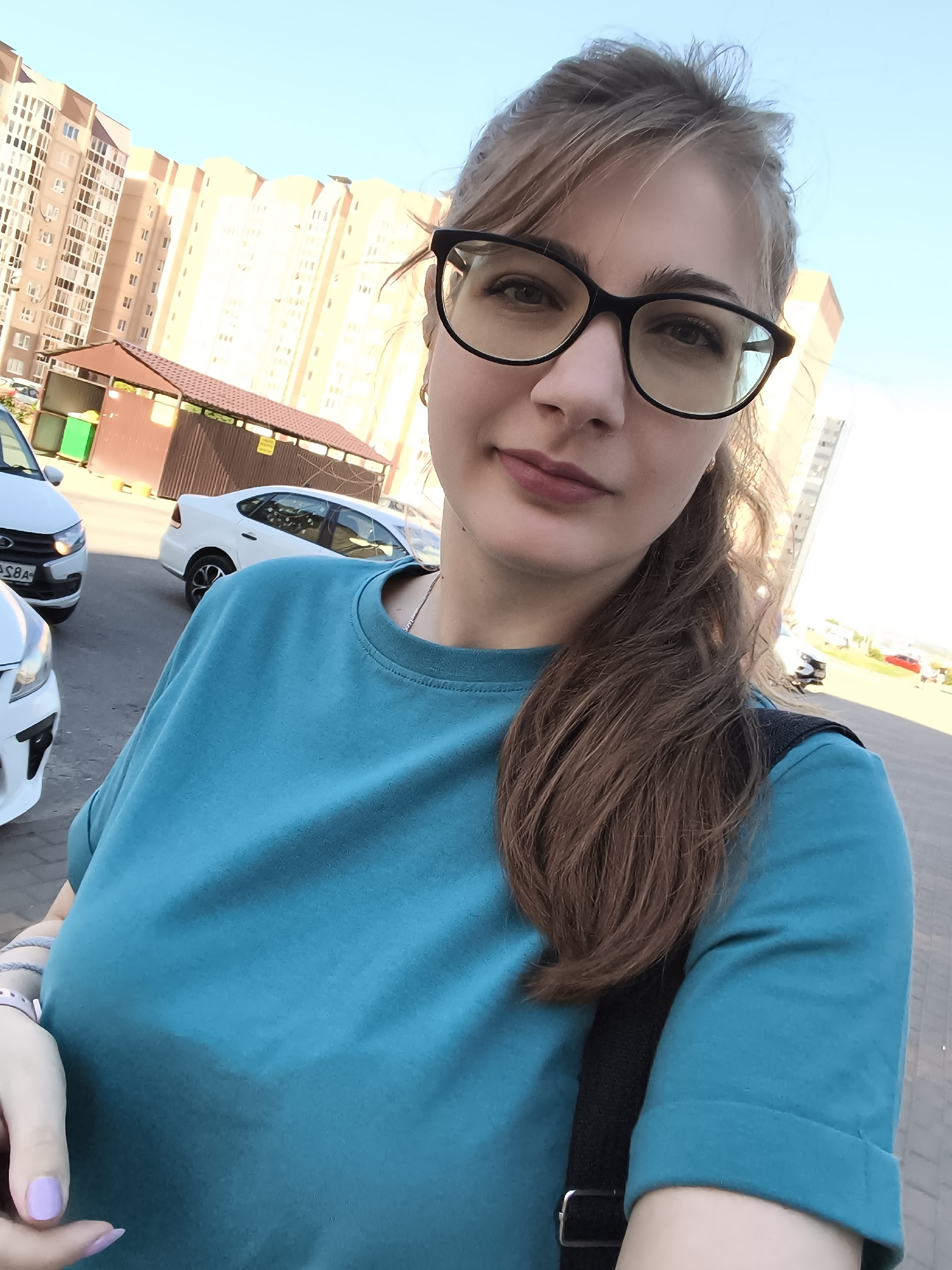 A young woman with brown hair and glasses is taking a selfie outdoors. She is wearing a blue t-shirt and a black backpack. The background shows a modern apartment building and parked cars. The sun is shining, indicating it is daytime. The woman has light skin and appears to be in a city or suburban area. She has light purple nail polish on her fingernails. There is no visible text in the image.