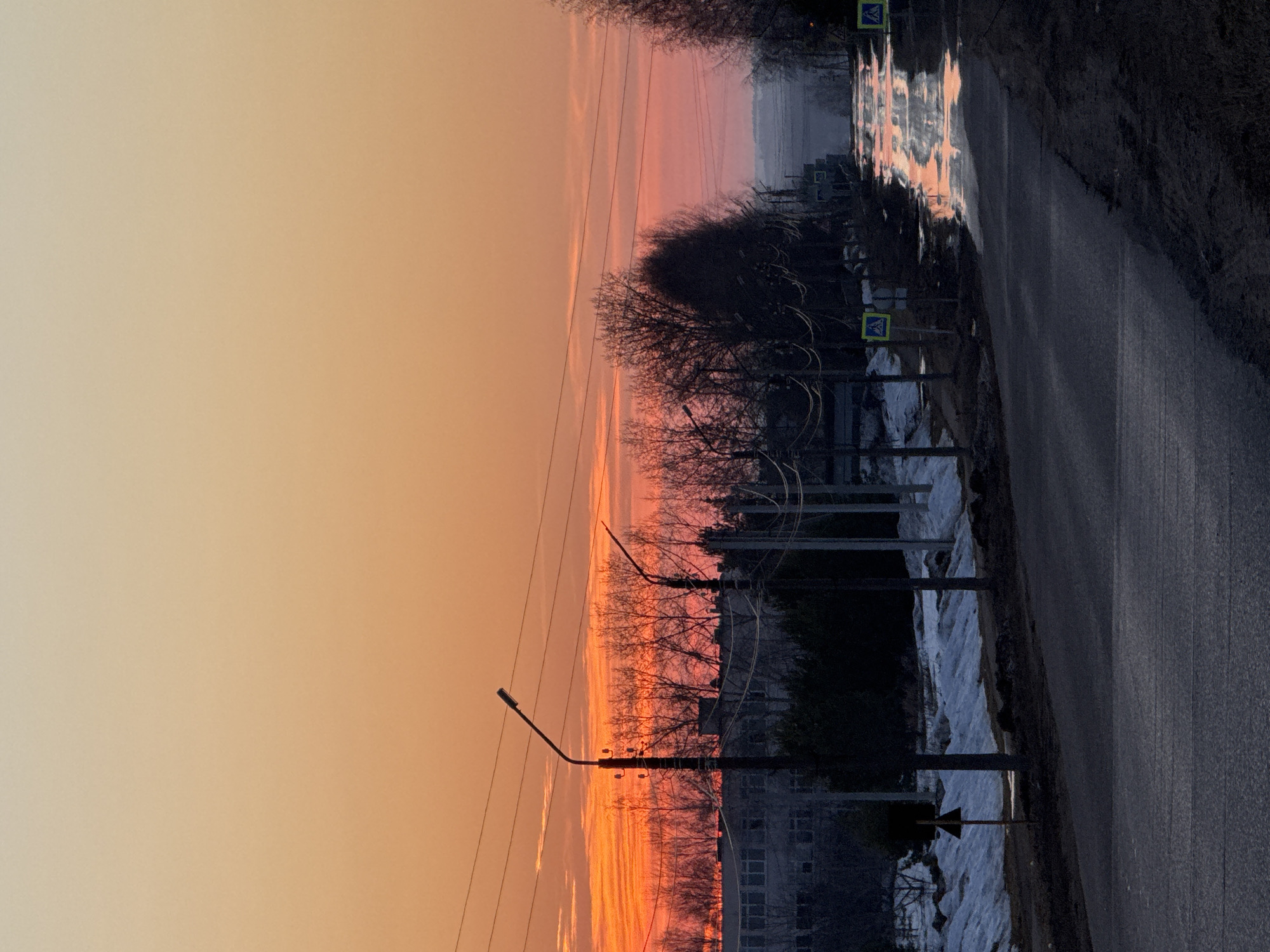 The photograph depicts a street scene in Прикамский, Russia at twilight. The sky transitions from orange to pink. A wet asphalt road stretches to the right edge. Streetlights and power lines run along the street's left side. Bare trees are visible between the streetlights and buildings. Patches of melting snow line the street. Pedestrian crossing signs are posted on poles. The image is slightly tilted, making the perspective appear distorted.