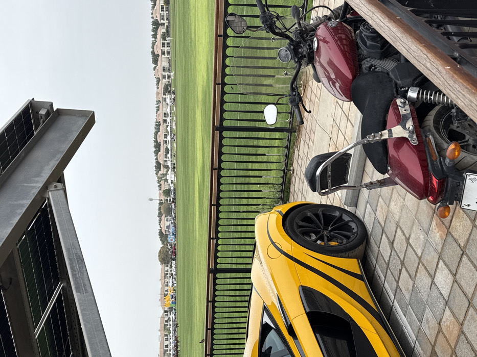An exterior scene depicts a bright yellow McLaren sports car and a dark red Harley-Davidson motorcycle parked on a light-colored paved block surface in the foreground, under an overhead structure fitted with solar panels. A black metal fence with a wooden top rail separates this area from a large, well-maintained green grass polo field.
In the mid-ground, on the polo field, several horses with riders are visible on the far left, suggesting ongoing or imminent equestrian activity. Various white tents and several parked vehicles are distributed across the field, with some distinct yellow and blue structures present, indicating an event setup. A small dark bird is present on the grass near the fence in the left-center.
The background features a long row of beige-colored residential villas with reddish roofs extending across the horizon, characteristic of upscale residential communities. A tall, slender communication tower stands in the mid-right background. The sky is uniformly overcast and light gray.
The location is identified as the **Al Habtoor Polo Resort & Club** in Dubailand, Dubai, United Arab Emirates, based on the specific architecture of the villas, the presence of a polo field, and the luxury vehicles, all consistent with the resort's offerings.