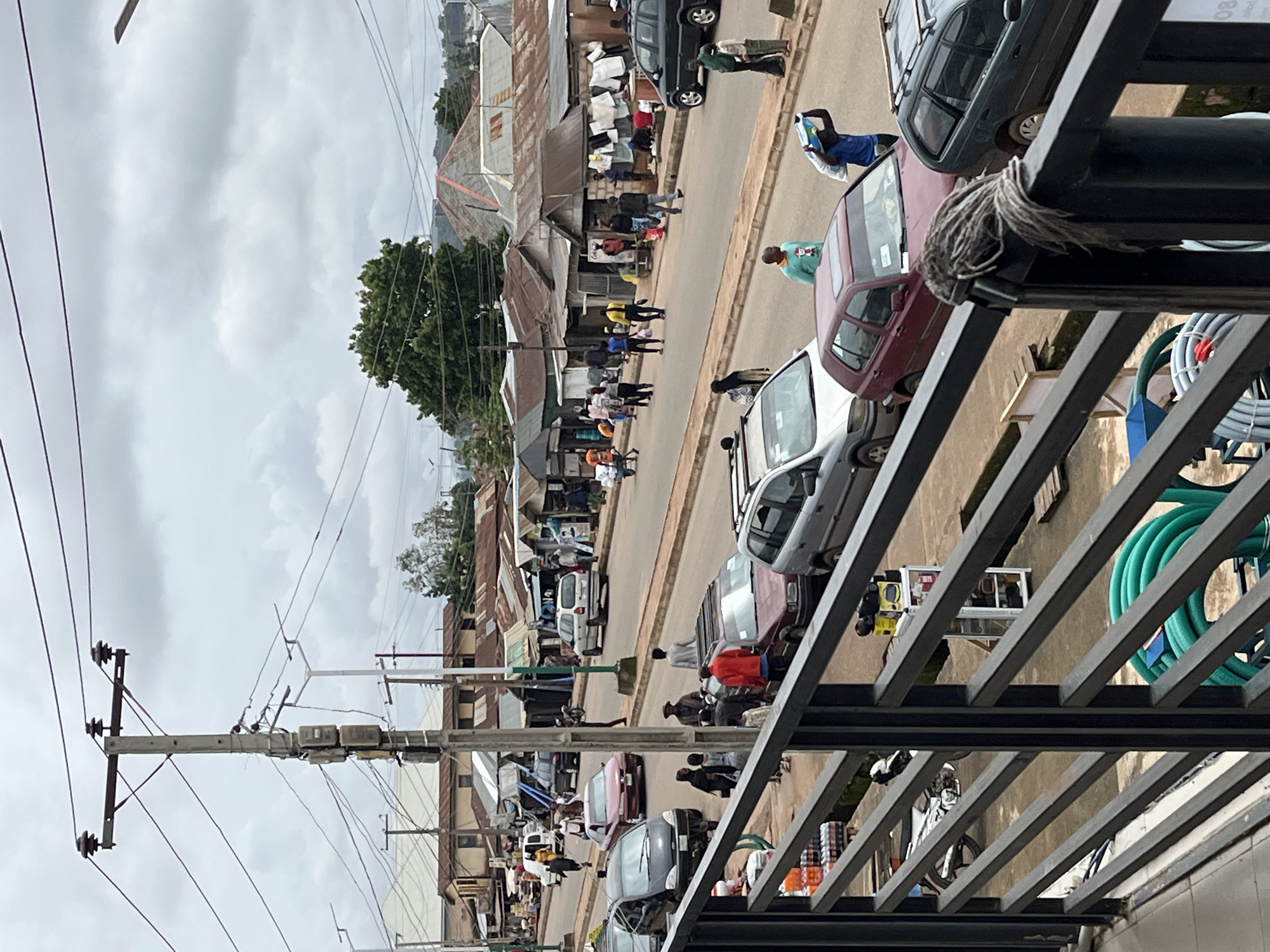 The image shows a street scene in Bukuru, Nigeria, under an overcast sky. The street is lined with buildings that appear to be shops or market stalls with corrugated metal roofs. Various vehicles, including cars and motorcycles, are present on the road. Several people are walking on the street and sidewalks, some carrying items. In the foreground, a black metal railing runs across the frame, partially obscuring the view of a motorcycle parked on the left and a coiled green hose on the right. There are also bottles of beverages stacked on shelves behind the railing. The overall impression is that of a busy marketplace or commercial area.