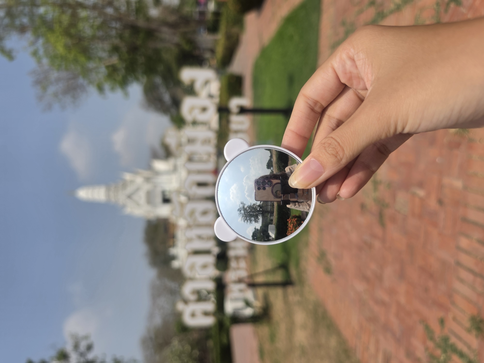 A person holds a white, circular mirror with bear ears in front of a white Thai temple and large Thai script sign in the Historic City of Ayutthaya, Thailand. The mirror reflects a person with a cell phone taking the photo. The foreground is a red brick pathway and green grass. The weather appears sunny with a few clouds.