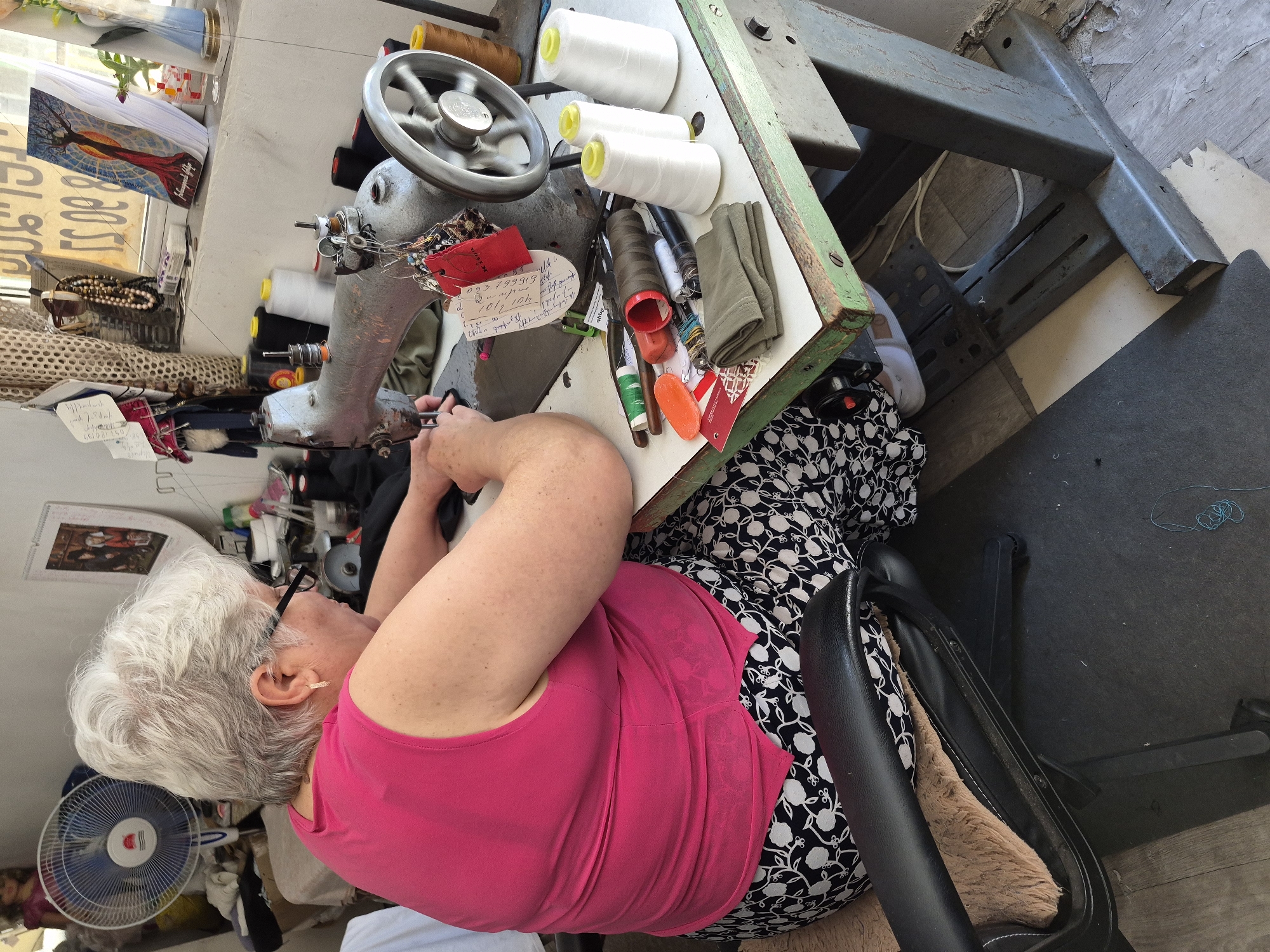 An overhead shot captures an elderly woman with short, wavy white hair diligently working at an industrial sewing machine in what appears to be a small tailoring workshop in Yerevan, Armenia. She is seated in a black office chair with a torn cushion, wearing a bright pink sleeveless top and a black-and-white patterned skirt. Her arms are bent over the machine, suggesting she is guiding fabric or making adjustments.
The sturdy, grey metal sewing machine dominates the foreground, its handwheel slightly blurred, indicating motion. The machine is set into an old, green-edged wooden tabletop, which is cluttered with numerous sewing essentials. White, brown, black, and green spools of thread are scattered across the surface, alongside various tools like scissors, pins, an orange bar of soap (possibly used as tailor's chalk), and measuring tapes. A red tag with "JISON" and a white tag displaying the phone number "093 799919" and a date "20/07/2021" are visible near the machine's needle.
The setting appears functional and well-used. Natural light streams in from a window on the left, where a reverse "80" is visible, likely from an external sign. Below the window, shelves hold more sewing items, including beads and fabric scraps. A small, white electric fan with a blue grate and "RED" branding is positioned on the floor near the woman, suggesting warm temperatures. The floor is covered in a mix of wooden panels and a dark mat. Peeling paint on a wall behind the machine adds to the shop's rustic character. Another framed image or calendar depicting a stylized tree with a red skirt, possibly labeled "Hagop Hago," hangs on the wall further back, alongside a paper with "100%". The overall scene depicts a moment of focused craftsmanship in a busy, lived-in workspace.
