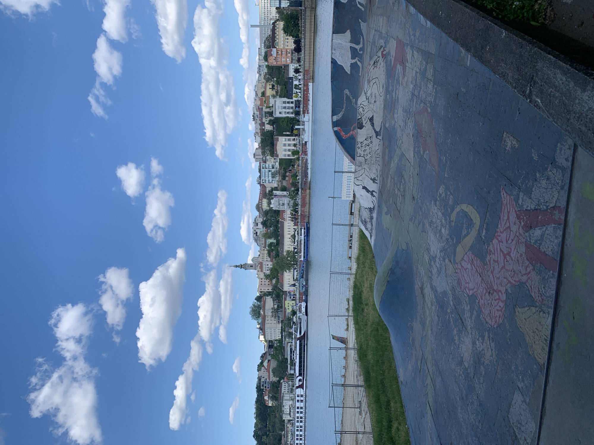 This image captures a vibrant, panoramic view of a city waterfront, identified as Belgrade, Serbia, on a bright, clear day. The scene is bisected by a wide river, likely the Danube or Sava, which flows between an urban skyline on the far bank and a decorative promenade in the foreground.
On the far side of the river, a dense collection of buildings forms the city's profile, ascending a gentle slope. The architecture is diverse, featuring multi-story residential and commercial structures with varied facades and rooftop styles, characteristic of European cities. A notable church spire rises above the buildings, adding a distinctive landmark to the urban landscape. Several large boats, including what appears to be a river cruise ship, are visible on the calm waters, either docked or navigating.
The sky above is a brilliant blue, liberally adorned with scattered, fluffy white cumulus clouds, indicating fair and sunny weather. In the foreground, on the right, there is a colorful, patterned walkway or wall, possibly a large mural made of tiles or painted concrete. This surface is decorated with various abstract and stylized shapes, figures, and patterns in shades of red, black, white, and yellow. Adjacent to this decorative surface is a strip of green grass, separated from the river by a fence or railing.
The overall atmosphere of the image is peaceful and scenic, showcasing a bustling urban environment under pleasant weather conditions. No legible text is visible in the image.