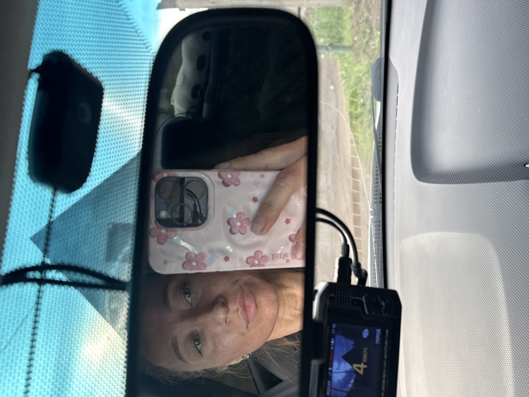 The image shows a rearview mirror selfie inside a car. The driver's face is visible in the mirror, holding a pink cell phone case with floral designs. A dashboard camera is mounted below the rearview mirror. The car is likely located in the city of Chaikovsky, Russia, based on the Cyrillic text ("ГОРОД") displayed on the dashboard camera screen. Outside the car, a road and some greenery are partially visible through the windshield. The car's dashboard is visible to the right of the mirror and a portion of the windshield is visible above the mirror.