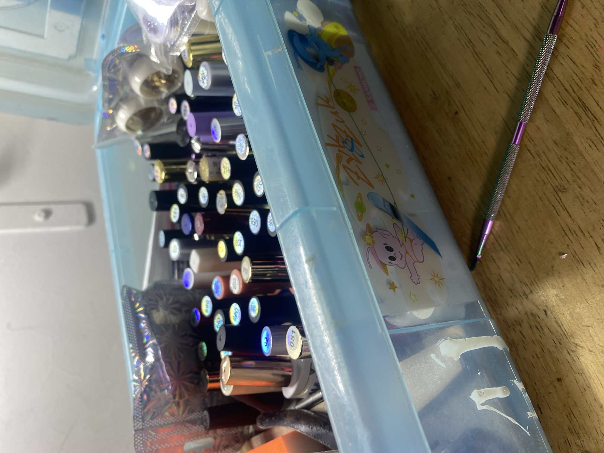 The image captures an angled, close-up view of a light blue plastic storage container filled with various nail art or beauty supplies, located in Cotoperíz, Venezuela. Inside the container, numerous cylindrical objects, likely gel polish bottles or tubes, are arranged horizontally. Many of these items have small, circular holographic stickers on their ends, displaying numbers such as 807, 010, 203, 890, and 051, indicating product codes. Small clear plastic bags containing glitter or decorative items are also visible among the contents.
Resting on a light brown wooden surface next to the container is a metallic, double-ended tool, possibly for nail art, featuring ridged handles and an iridescent purple color. Tucked inside the container, on the right side, is a piece of paper or card with colorful cartoon illustrations, including a pink, bunny-like character, stars, planets, and partially visible stylized text that appears to include "Art". The background on the left is a plain, light-colored surface. The scene depicts an organized collection of craft or cosmetic materials.