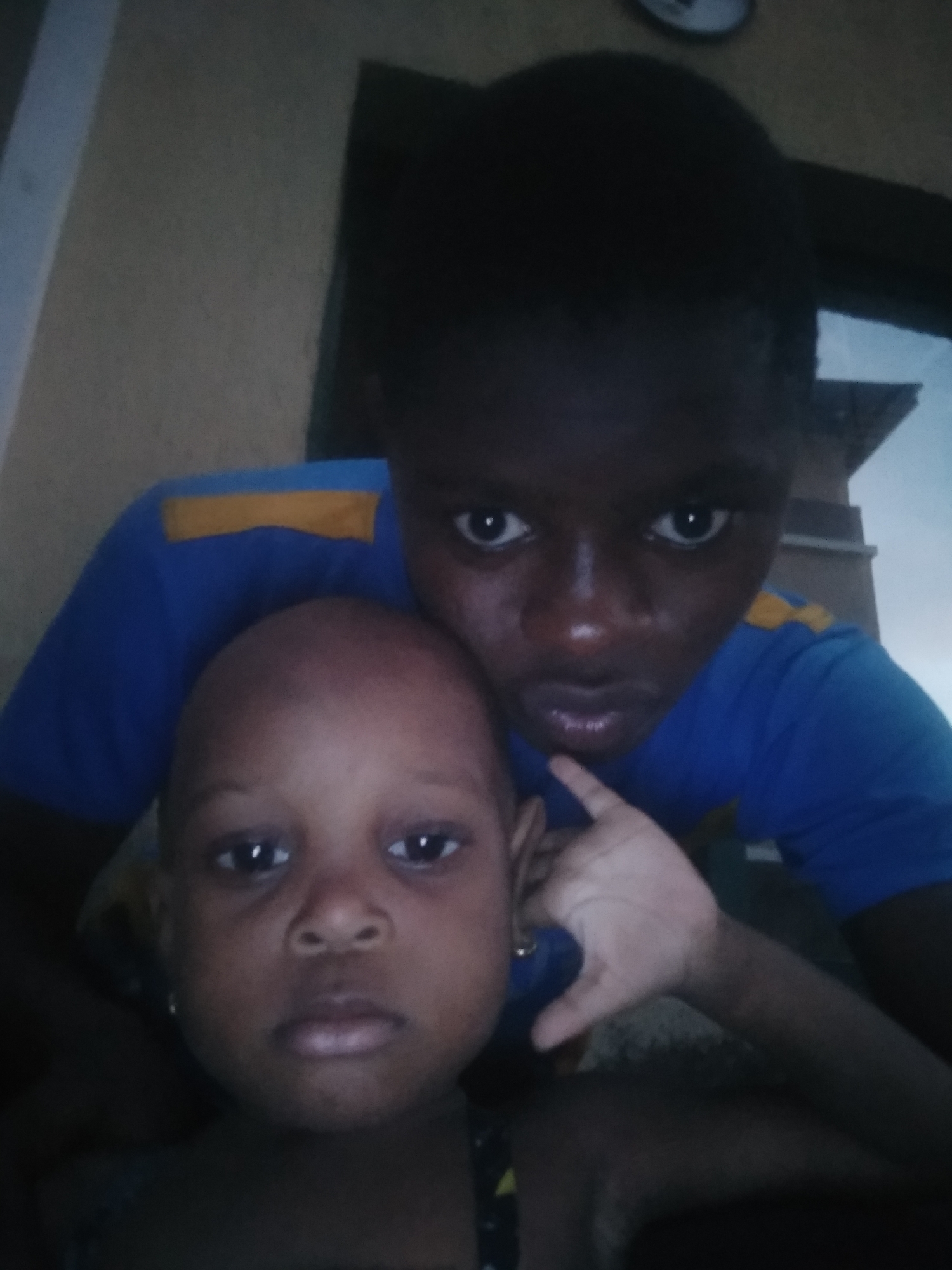 The image displays a close-up photograph of two individuals, likely of African descent, positioned indoors.
In the background, an older individual, appearing to be a teenager or young adult, is visible. They have dark skin and are wearing a blue long-sleeved shirt with a horizontal yellow stripe across the upper chest/shoulder area. Their dark eyes are wide open and looking directly at the camera, while their mouth is slightly pursed. Their head is positioned above and behind the younger child. A hand with a lighter skin tone, presumably belonging to this person, is visible near the younger child's right ear.
In the foreground, a younger child, possibly a toddler, is prominent. This child also has dark skin and a shaved head, with large, dark eyes looking straight at the camera. Their expression is neutral.
The setting appears to be an indoor space with a plain, light-colored wall forming the background. A circular clock is partially visible in the top center of the image, slightly out of focus. The overall lighting is somewhat dim, suggesting an indoor environment without strong natural light, possibly indicating evening or night. The individuals appear to be posing for a close-up photo or a selfie. There are no specific visual cues within the image to confirm the location, but the subjects' appearance is consistent with the provided context of Shangisha, Nigeria. No visible text is present in the image.