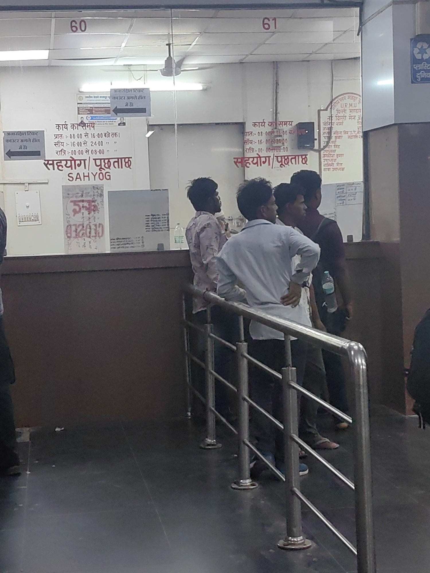 The image depicts a scene at a railway station ticket counter in Delhi, India. The prominent signage indicates "60" and "61" at the top, likely representing counter numbers. A sign in Hindi and English states "कार्य का समय" (Work Time) and lists operating hours for different shifts, including morning, afternoon, and night. Another sign with Hindi text and an arrow points to "सहयोग/पूछताछ" (Cooperation/Inquiry).
Several individuals are visible, appearing to be passengers or station staff. Three men are standing in the foreground, one in a light blue shirt and dark pants, another in a maroon shirt, and a third in a patterned shirt. They seem to be waiting or interacting near the counter. In the background, more people are partially visible.
The environment includes a counter area with glass partitions, illuminated by fluorescent lights. Various signs, posters, and what appear to be information displays are mounted on the wall behind the counter. A ceiling fan is also visible. The floor is dark and tiled, with some scattered debris. Metal railings are installed in the foreground, likely to manage queues. A blue plastic water bottle is placed on the counter. A sign with a recycling symbol and Hindi text about "Plastic" is mounted on the right side. The overall impression is of a functional but somewhat crowded public service area.
