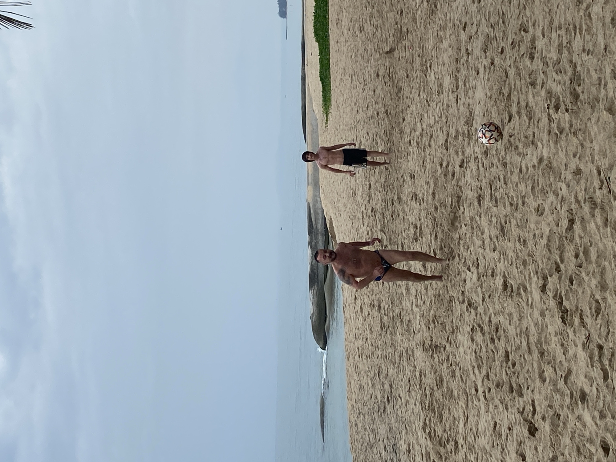 Two men are on a sandy beach playing with a soccer ball. A body of water is to the left of the frame. The beach is in Phu Thuy Ward, Vietnam. The sky is overcast. One man is wearing swimwear and another is wearing dark shorts.