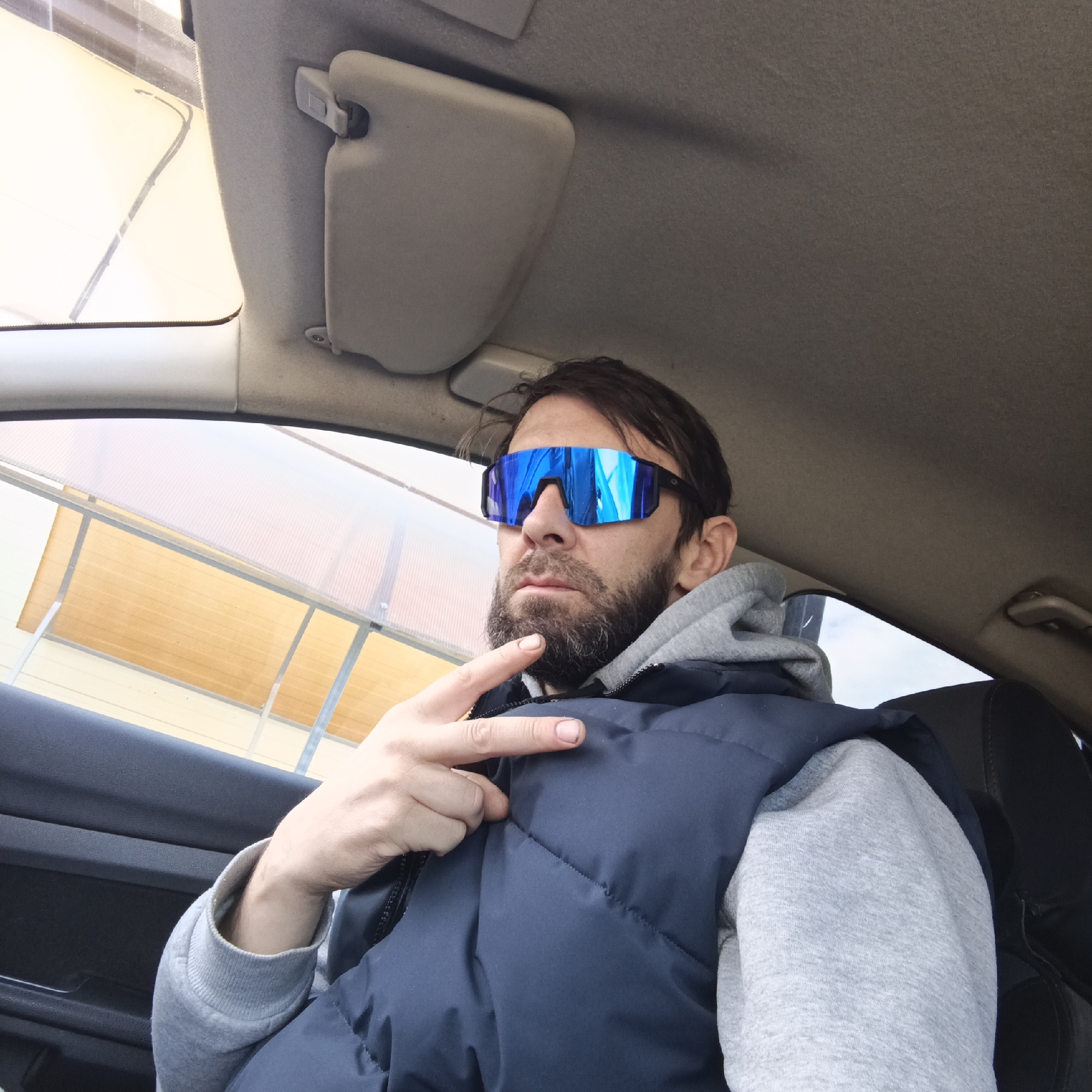 A man is depicted inside a car in Chaikovsky, Russia, taking a selfie or being photographed from a low angle. He occupies the driver's or passenger seat, looking slightly to his right. He wears a pair of striking blue reflective sunglasses, a gray hooded sweatshirt, and a dark blue puffer vest. He has dark hair and a beard. His left hand is raised, forming a peace sign.
The car's interior features prominently, including the light-colored headliner, a sun visor on the right, and parts of the dashboard and door panel. Bright, diffused daylight enters through the windows. Outside the front passenger window, a structure with a translucent, ribbed or corrugated yellow-orange roof or awning is visible, supported by metal beams. This suggests a covered outdoor area, possibly an entrance to a building or a car wash. The time of day appears to be daytime, with soft, indirect lighting. The man's expression is neutral due to the sunglasses, and his hand gesture conveys a casual or friendly demeanor. No visible text is present in the image.
