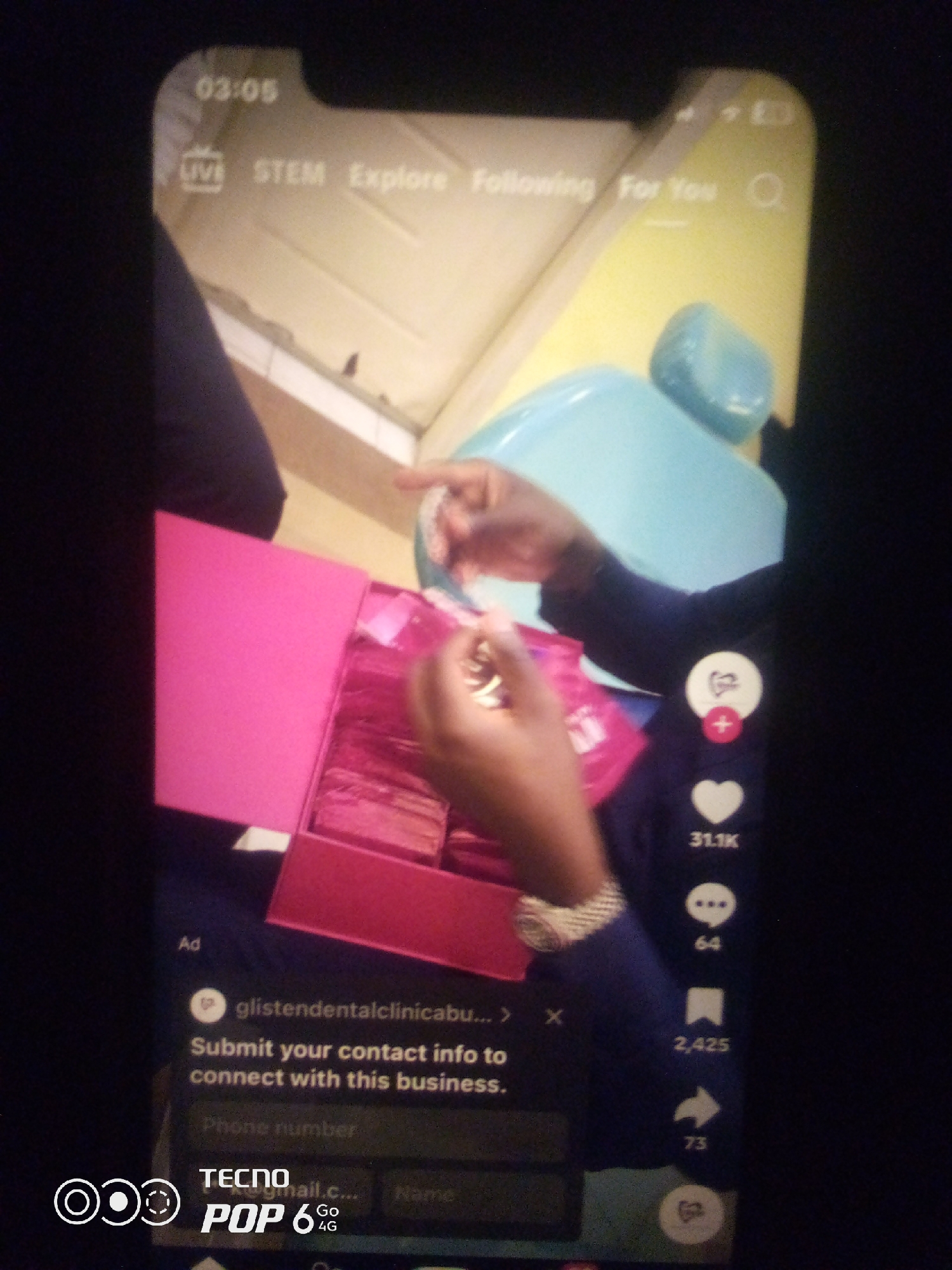 The image is a screenshot of a TikTok video. The video displays a pink box filled with individually wrapped items, possibly samples or products. A person's hands are visible interacting with the box, reaching in to pick up one of the items. The video interface shows standard TikTok elements: a timer at the top (03:05), navigation categories (LIVE, STEM, Explore, Following, For You), a search icon, and social interaction icons on the right side (heart with 31.1K likes, speech bubble with 64 comments, bookmark with 2,425 saves, and a share icon with 73 shares). Below the video content, there's an "Ad" section with text that reads "glistendentalclinica..., Submit your contact info to connect with this business." The bottom of the screen shows "TECNO POP 6 GO 4G" branding. The scene appears to be indoors, with a light yellow wall and a light blue object in the background. The image does not contain any specific visual cues to determine the location within the city of Howayi, Nigeria.