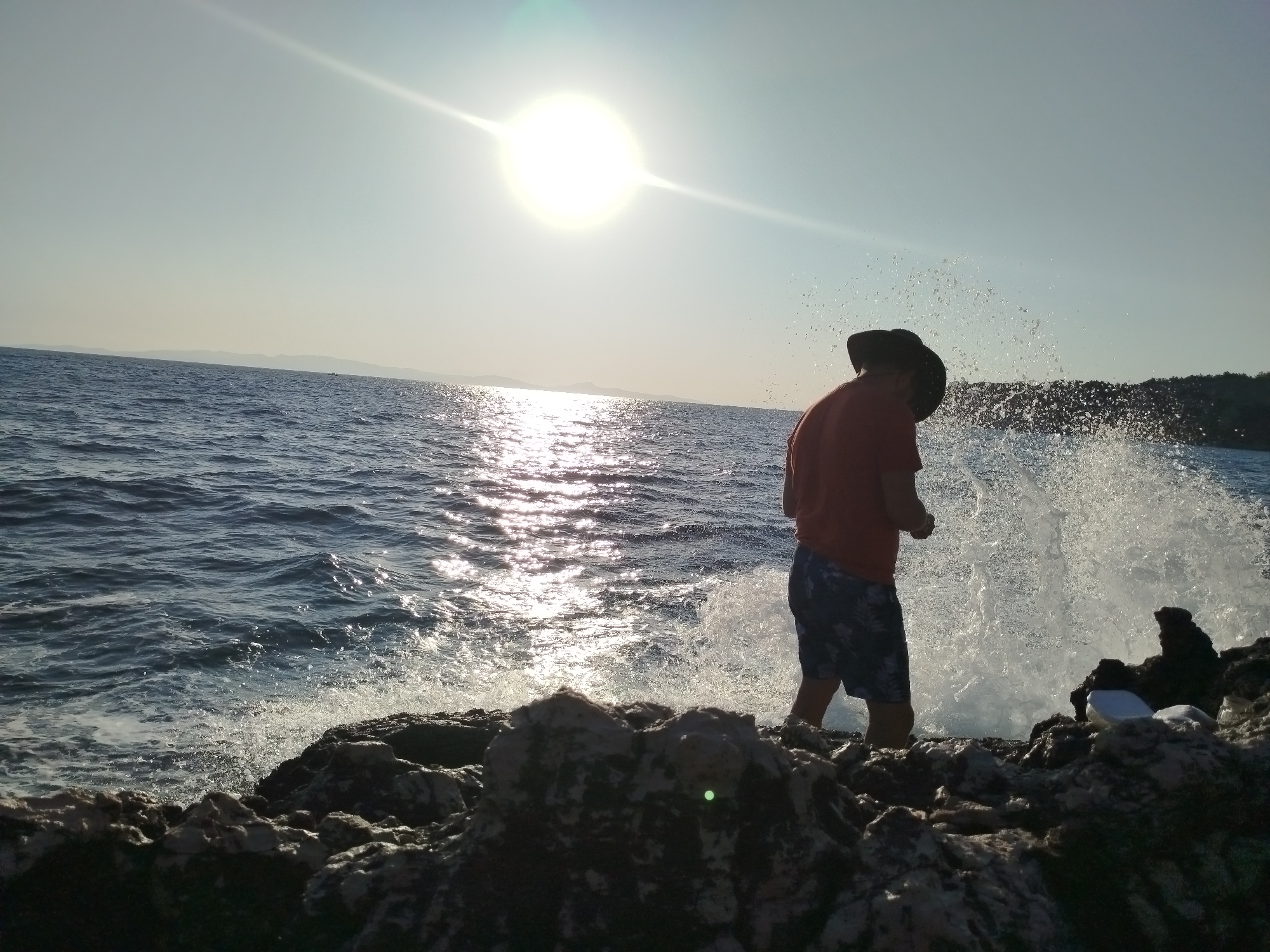 A person wearing a red shirt, dark shorts, and a dark hat stands on a rocky shoreline, silhouetted against the bright sun. The sun is high in the sky, creating a strong lens flare. Waves crash against the rocks, sending spray into the air. The water is a deep blue, and the sky is a pale blue. In the distance, there is a hazy coastline with hills. The scene appears to be in Karaburun, Turkey, likely during the day.