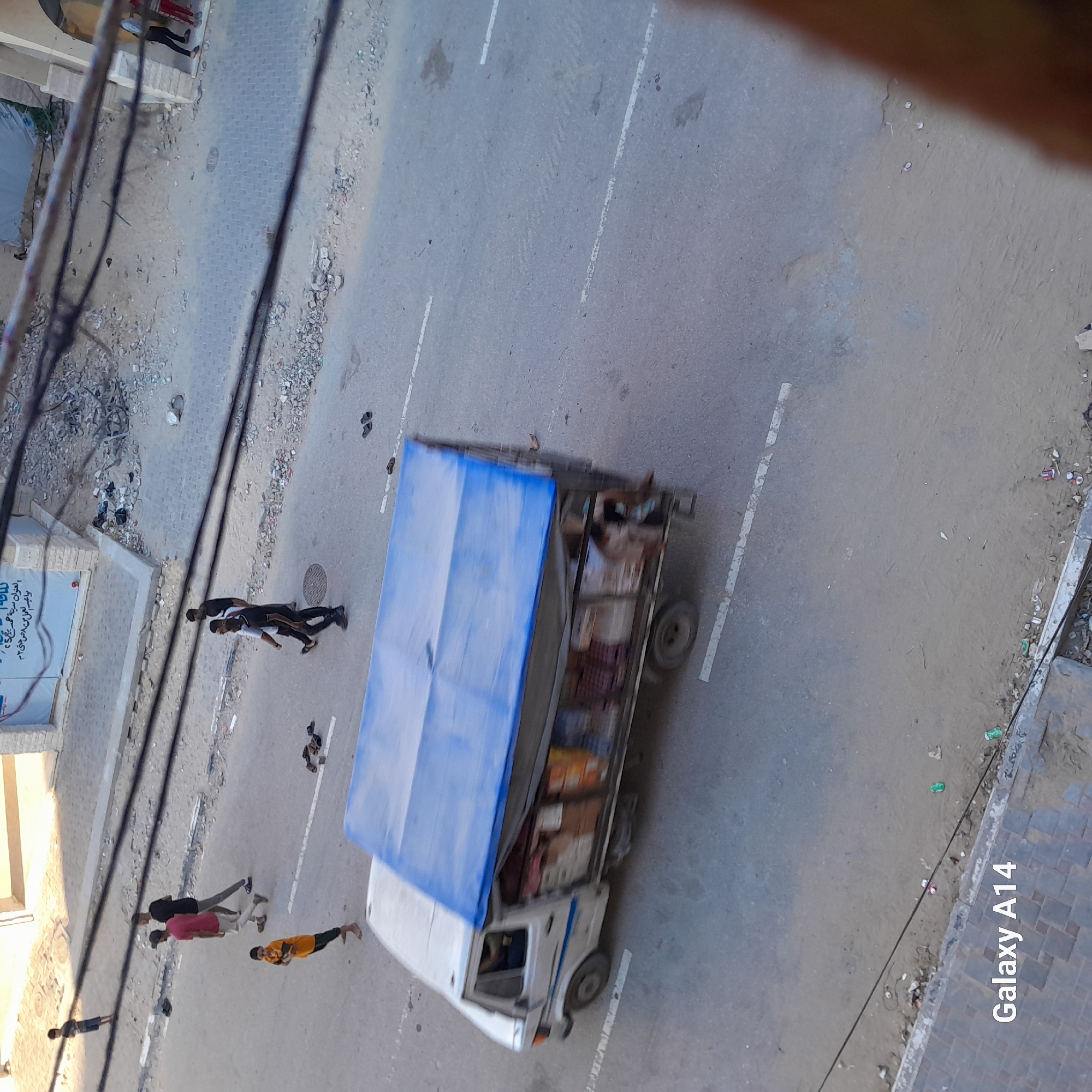 In Khan Yunis, Palestinian Territories, an aerial view captures a street scene. A white truck with a blue tarp-covered bed, filled with various boxes and items, is driving on the road. Several individuals are present on the street and sidewalk. Two young boys are walking on the road, one in an orange shirt and the other in a dark shirt and shorts. Further down the road, three young men are walking together, one in a pink shirt, one in a black t-shirt, and another in a black shirt and pants. Several pairs of sandals are scattered on the asphalt. In the background, a damaged building with exposed rebar and rubble suggests recent conflict or destruction. The environment appears to be an urban area with a paved road, a sidewalk, and debris.