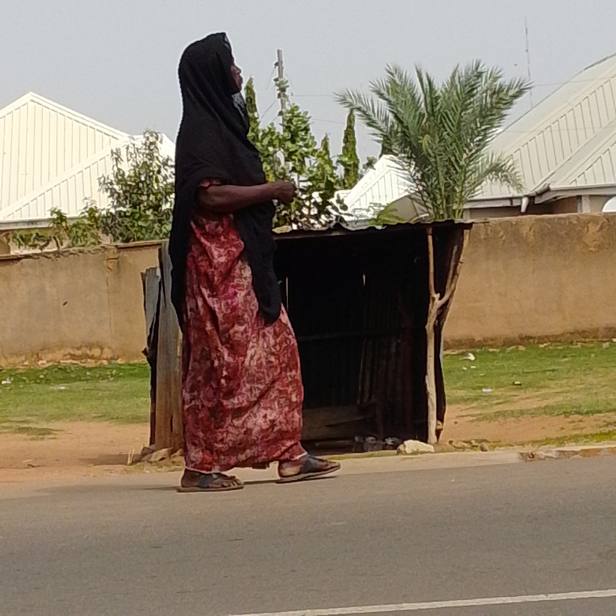 A lone woman, wearing a black hijab and a red floral patterned dress, walks along a paved road. She is viewed from the side, in profile, facing left and slightly away from the camera. Her feet are clad in dark sandals. She walks past a dilapidated, dark-colored stall or shelter constructed with corrugated metal sheeting and wooden supports. In the background, there is a light-colored, sloped roof of a building and a palm tree. Further in the distance, a brown wall borders a patch of green grass. The scene appears to be an outdoor urban environment, possibly within Gombe city, Nigeria, given the visual cues of the architecture and vegetation. The lighting suggests it is daytime.