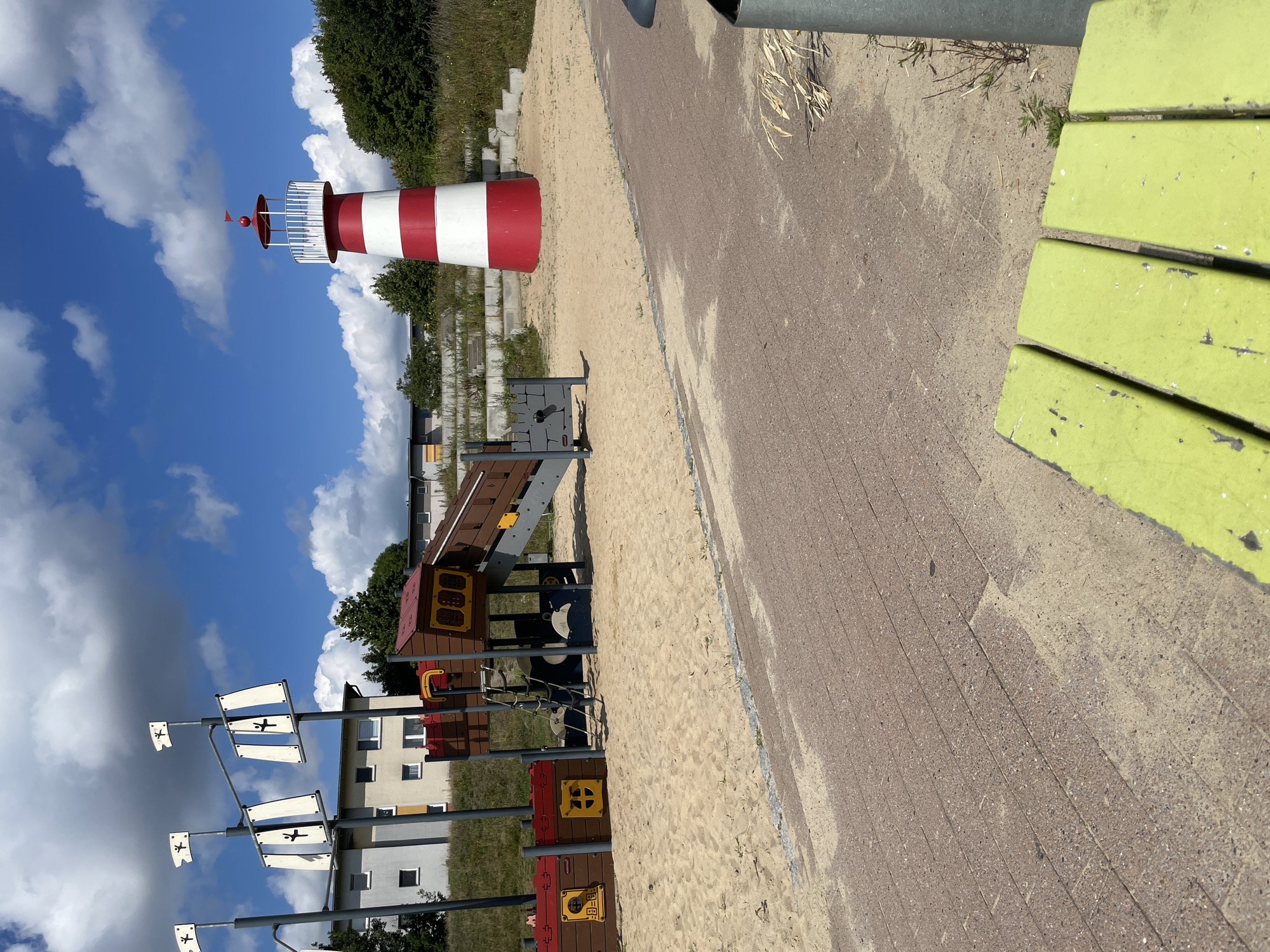 The image depicts a playground with a maritime theme on a sunny day. In the background, a red and white striped lighthouse stands prominently, a common visual element in coastal towns. Adjacent to it, a multi-level play structure resembling a pirate ship is visible, complete with masts and flags. The playground area is covered in sand, with a paved pathway running through the foreground. In the distance, apartment buildings can be seen, suggesting an urban or suburban setting. The sky is blue with scattered white clouds. This scene is consistent with a public park or recreation area within Bremerhaven, Germany, a city known for its port and maritime history.