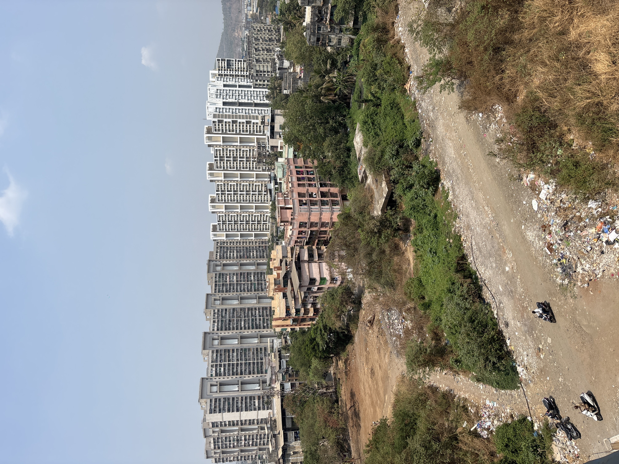 The image depicts a view in Ambernath, India, showing a juxtaposition of urban development and environmental degradation. On the left side, there are rows of tall, modern apartment buildings under a light blue sky. Moving rightward, the buildings transition to older, less maintained structures. A large area of barren land divides the buildings from a dirt path littered with garbage. Two motorcycles are parked on the path, and one person is riding a motorcycle. The path is bordered by green vegetation. The garbage, barren land, and the presence of the motorcycles indicate human activity and the environmental impact associated with it. In the background, there is a line of hills/mountains.