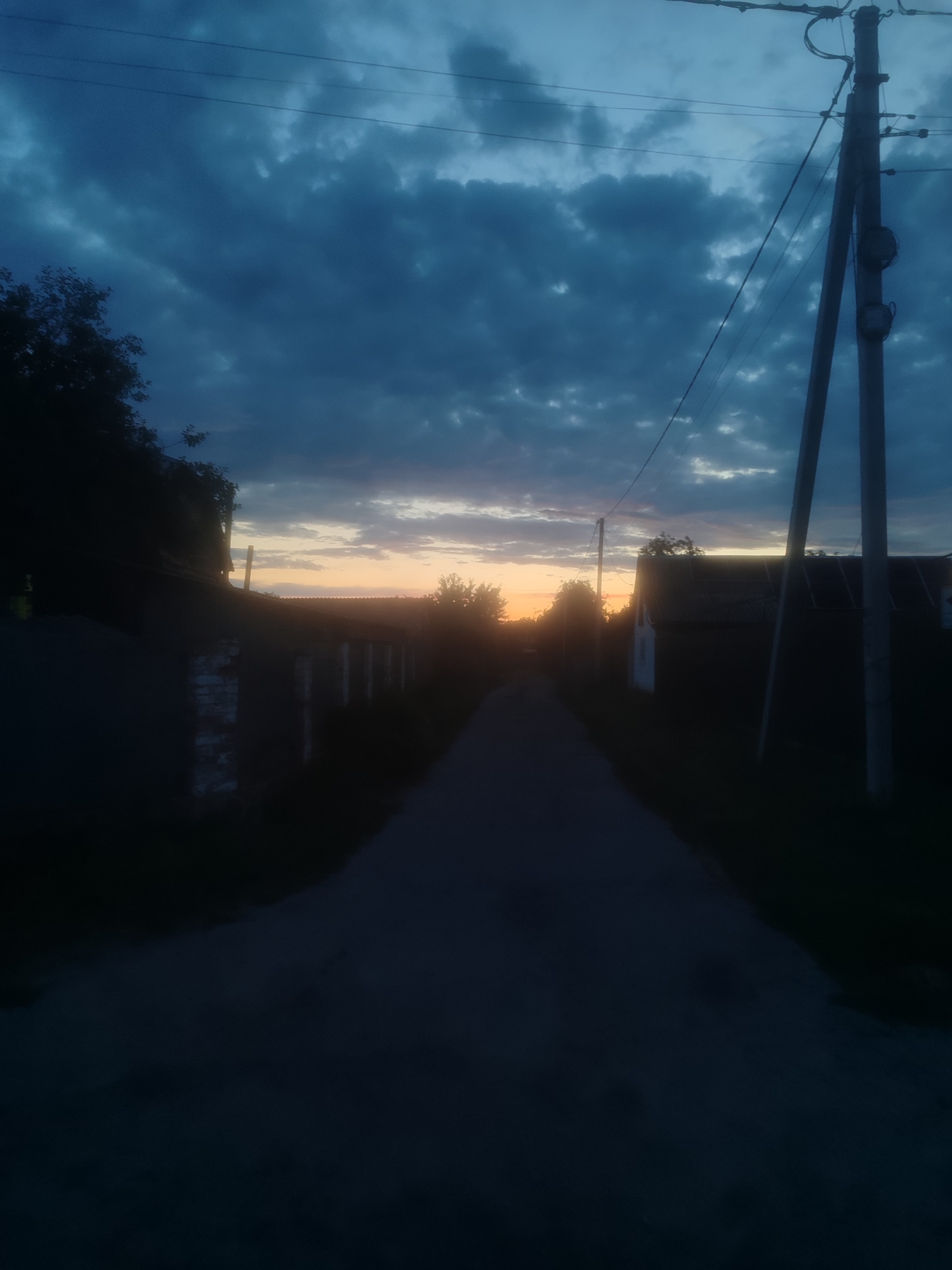 The image depicts a rural road at dusk, with a dramatic sky filled with dark, heavy clouds. A faint orange glow is visible on the horizon, suggesting the sun is setting. On either side of the road, shadowy structures and trees are visible. A power line pole with wires is prominent on the right side of the image. The overall mood is somber and still. The scene is consistent with a rural or suburban area outside of a major city.